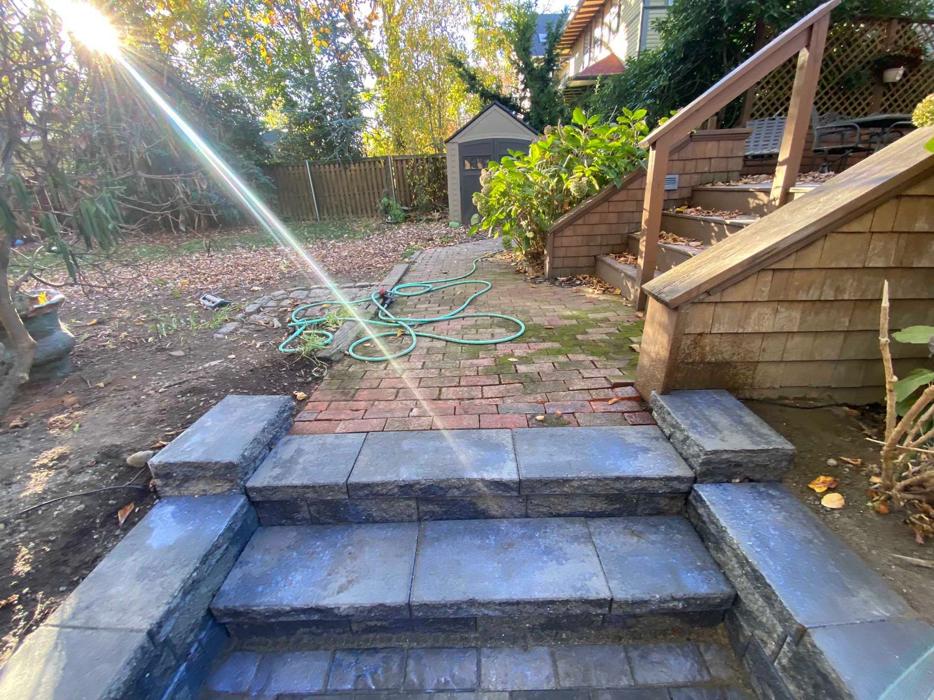 New stone steps lead to a brick path in a backyard with wooden stairs and a small shed in the background.