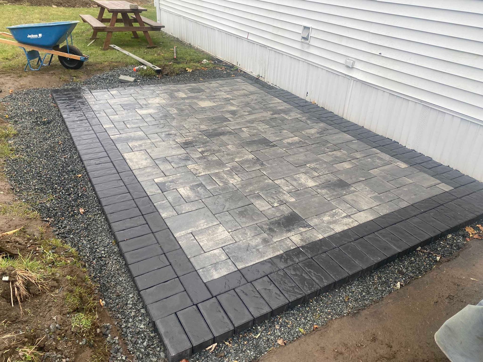 A rectangular stone paver patio with a dark border installed alongside a house, with gravel edging and a wheelbarrow nearby.