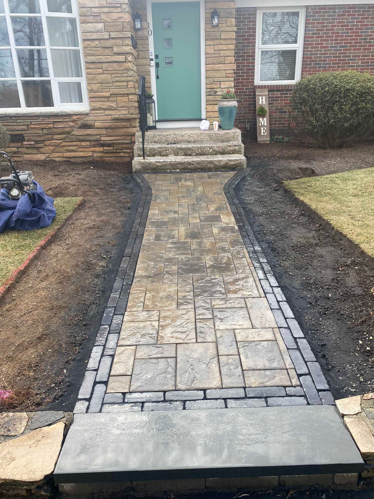 A newly installed stone paver walkway with a dark border leads to the concrete steps and teal front door of a home.