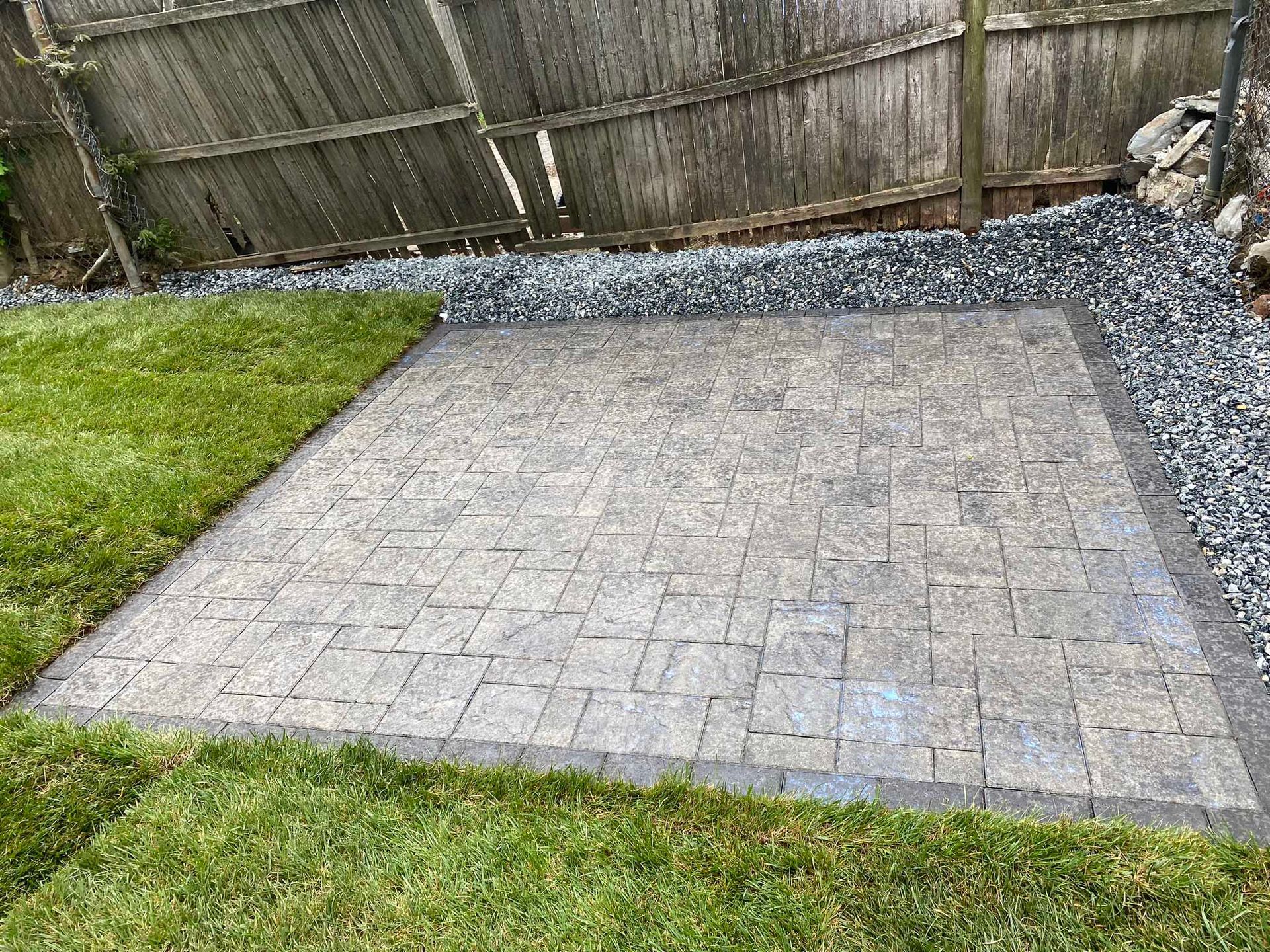 A rectangular patio made of grey pavers, surrounded by gravel and green grass, set against a wooden fence.