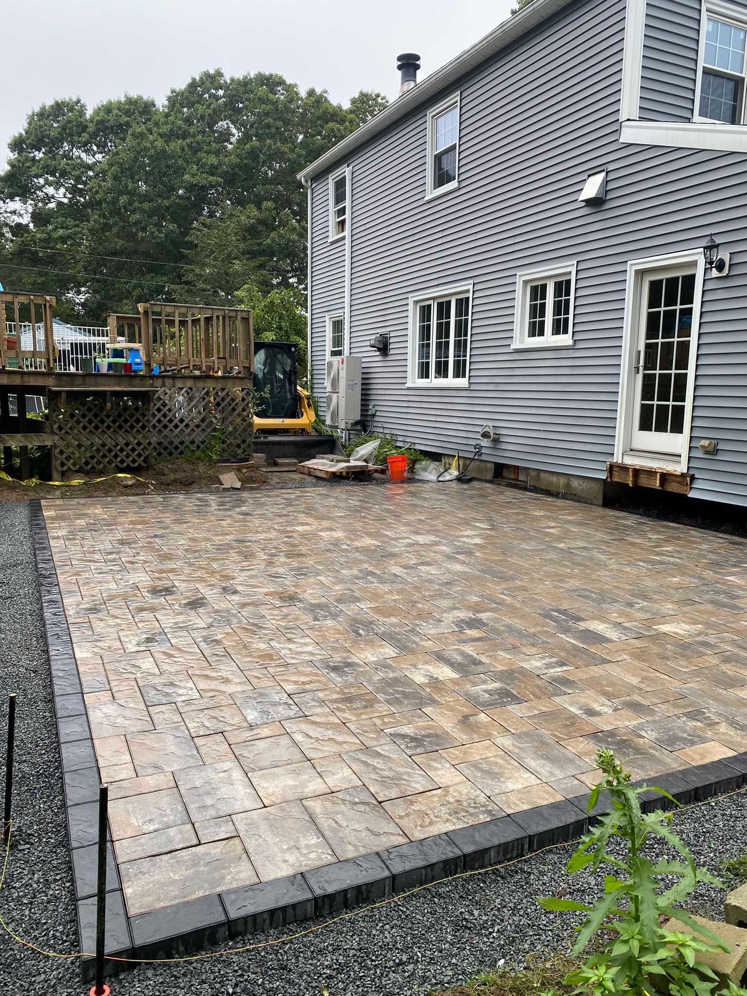 A newly installed stone paver patio sits against the side of a grey house with a wooden deck nearby.