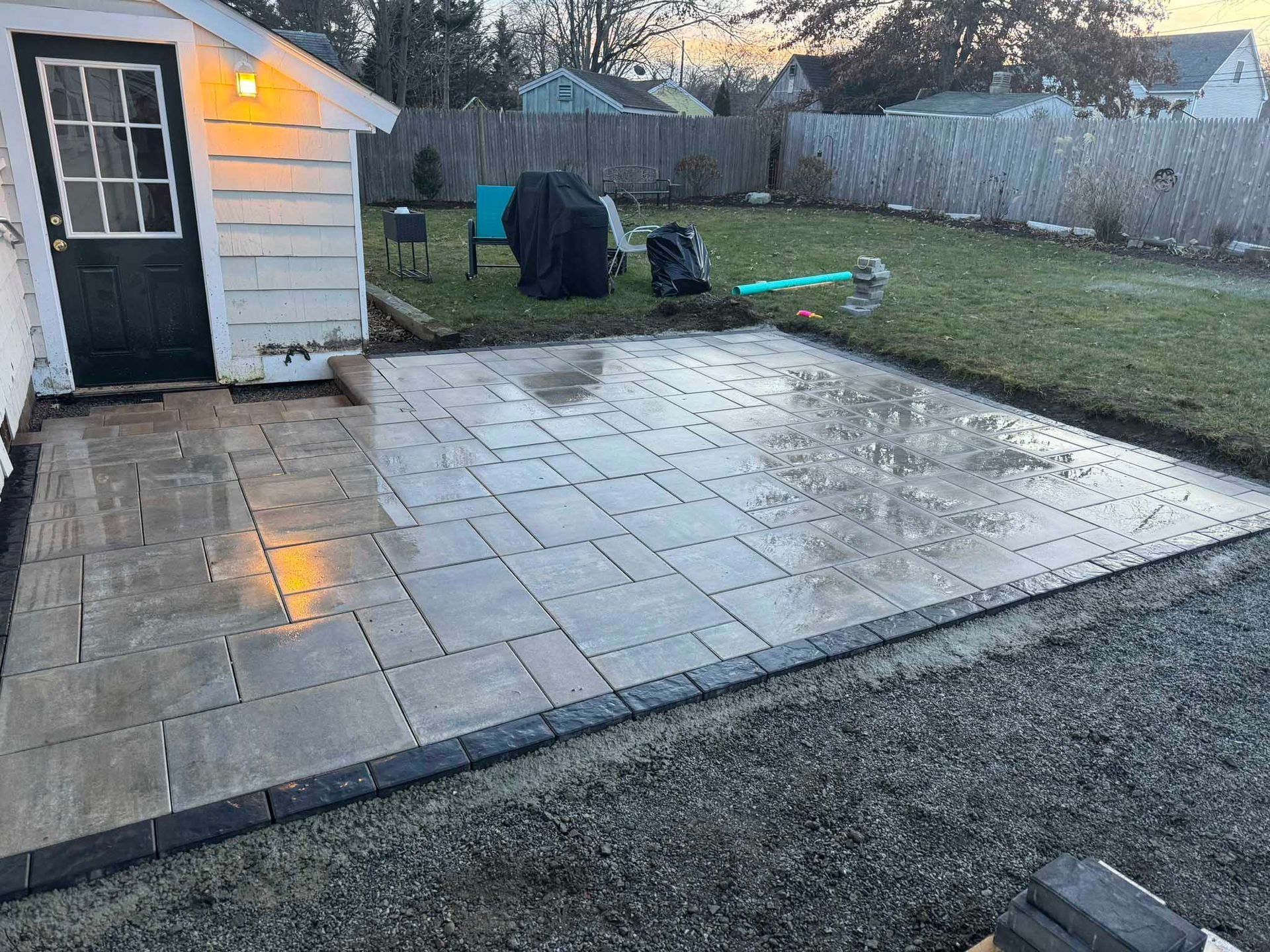A newly installed paver patio with a dark stone border, located next to a white shed in a backyard with a grass lawn.
