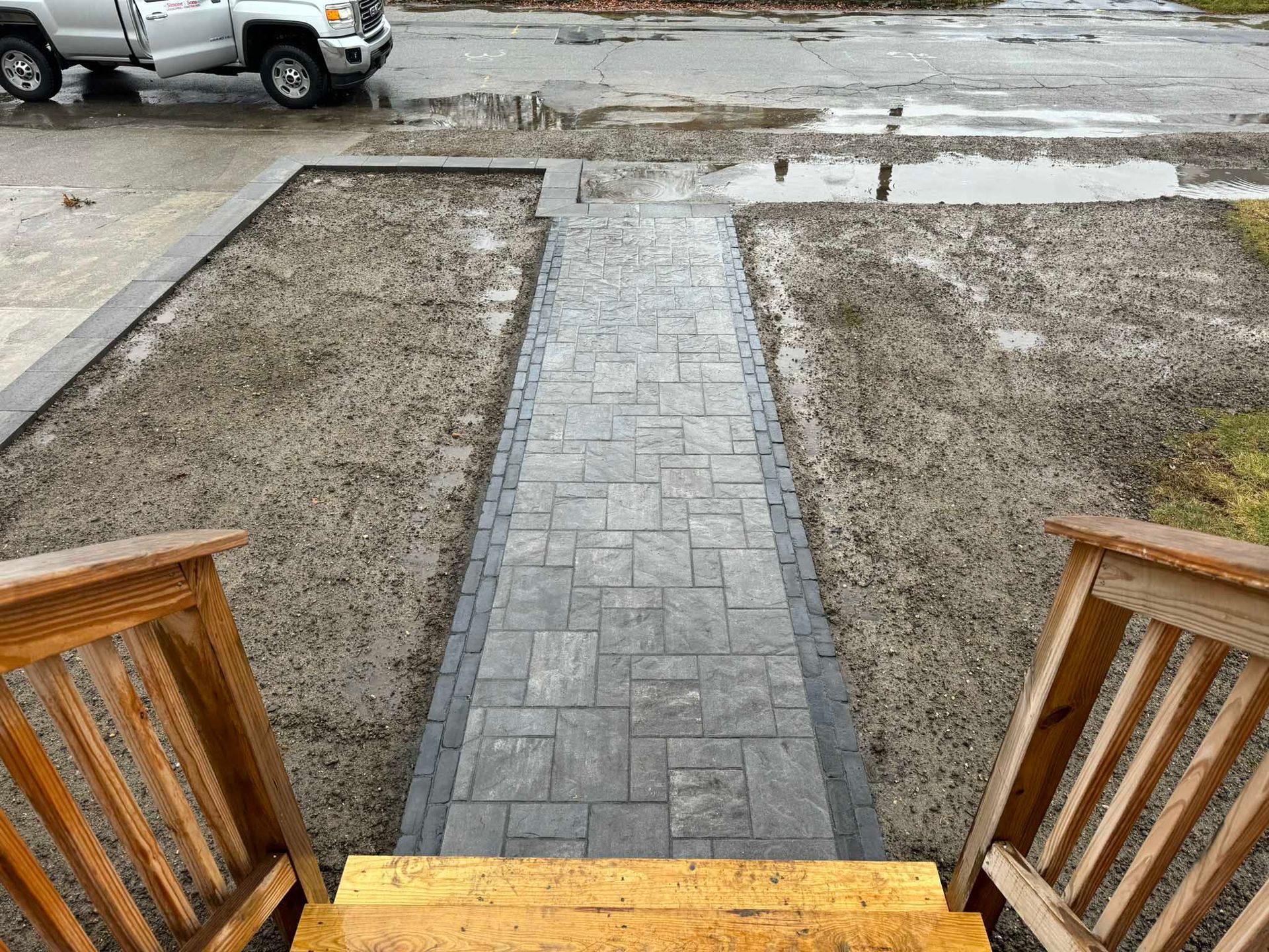 A new gray stone paver walkway extends from wooden porch stairs toward a road, flanked by unfinished dirt yards.