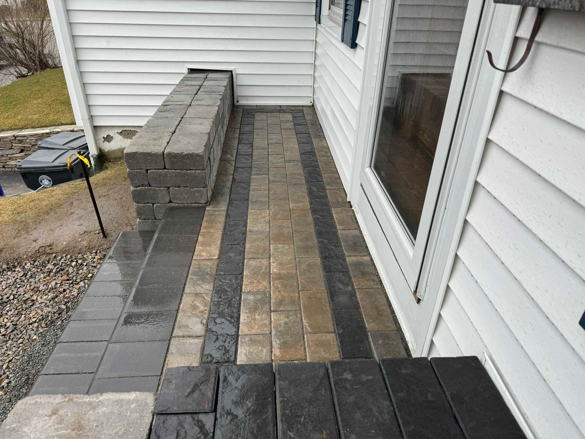 A paved walkway with light and dark stone rows leads to a home entrance, next to a matching low stone bench.