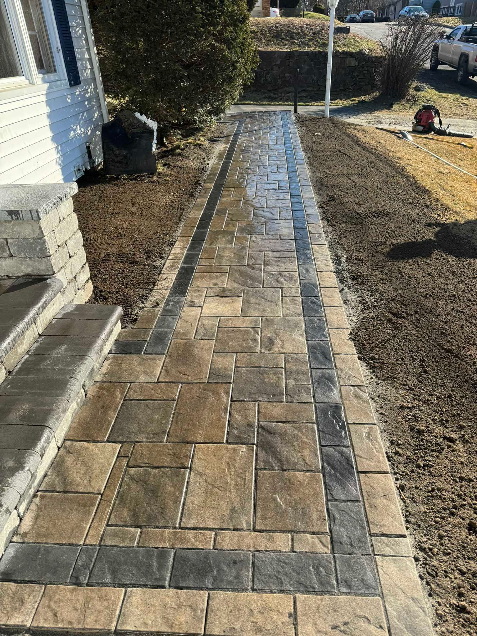 A new paver walkway with a dark grey border and tan, multi-sized stone center leading from a house to a street.
