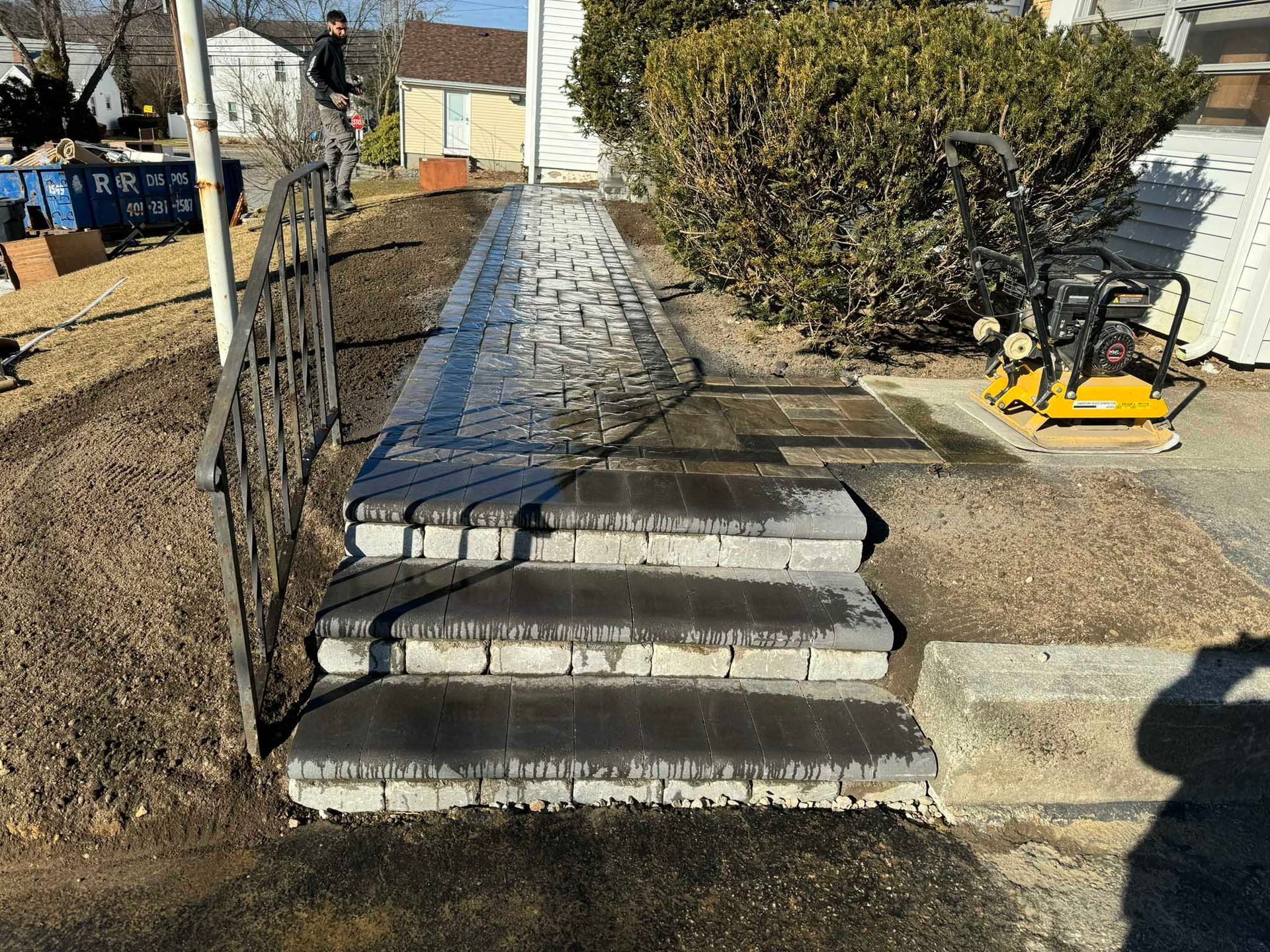 New dark brick walkway with tiered steps leading to a house entrance, with construction equipment nearby.