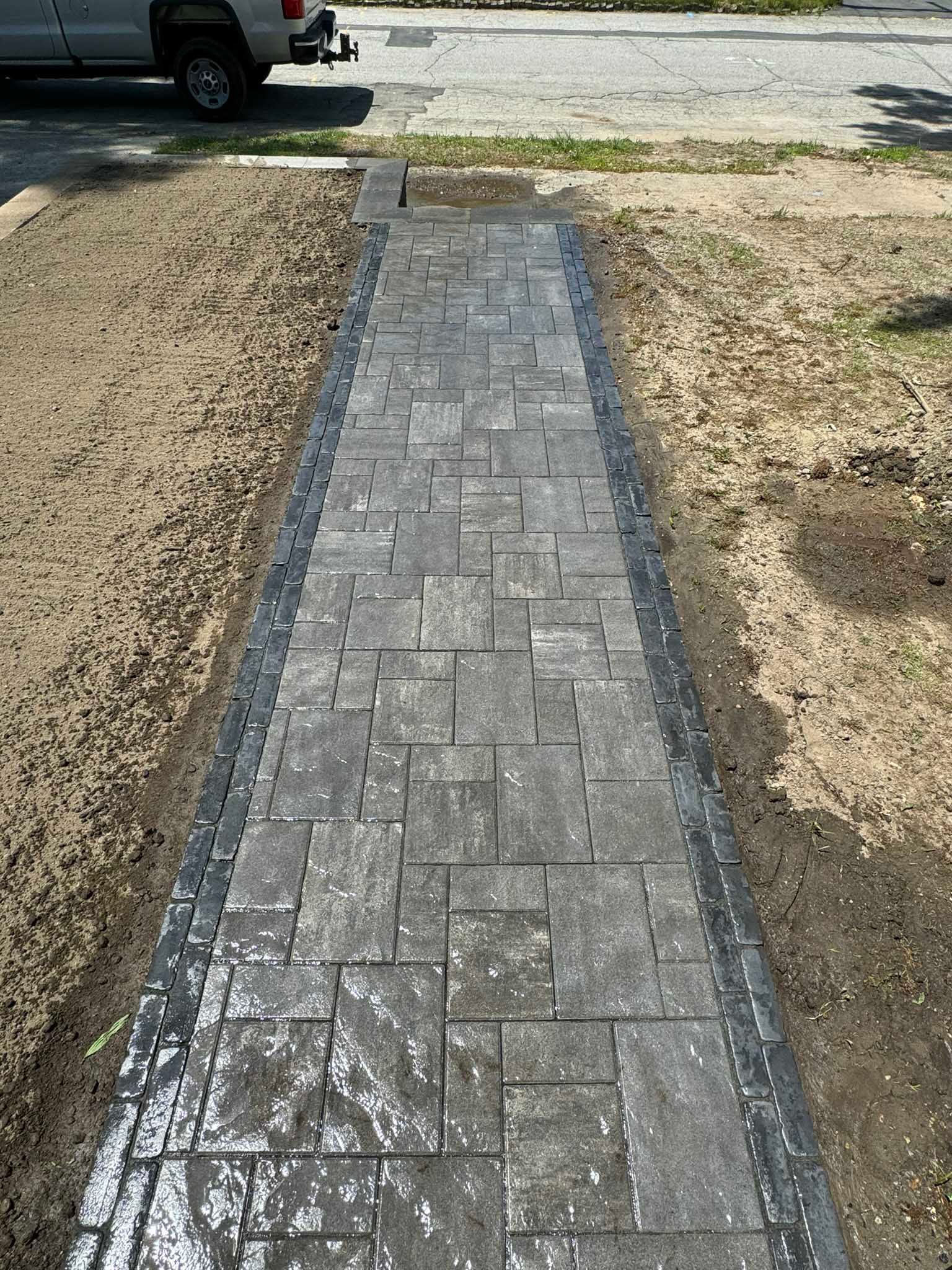 A newly installed gray paver walkway with a dark border, flanked by dirt patches and a truck in the background.
