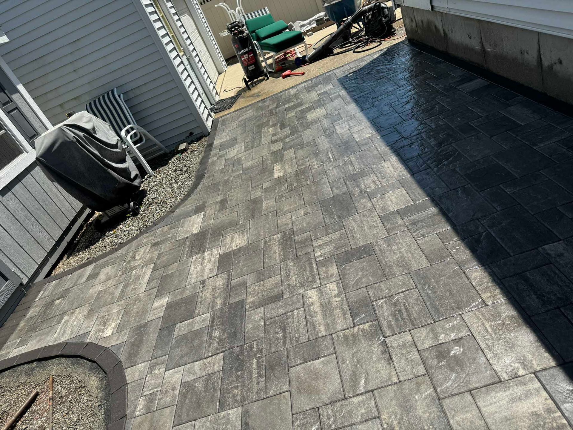 A gray-toned paver patio extending from a house, featuring a curved border and a wet, darker section near the foundation.