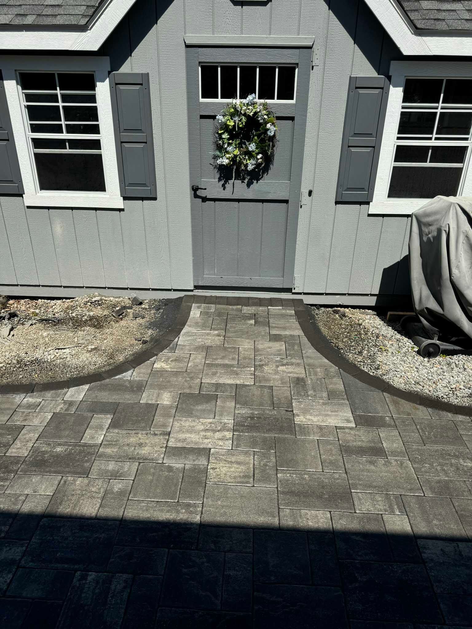 A gray wooden shed with a wreath on the door and a stone paver walkway leading to the entrance.
