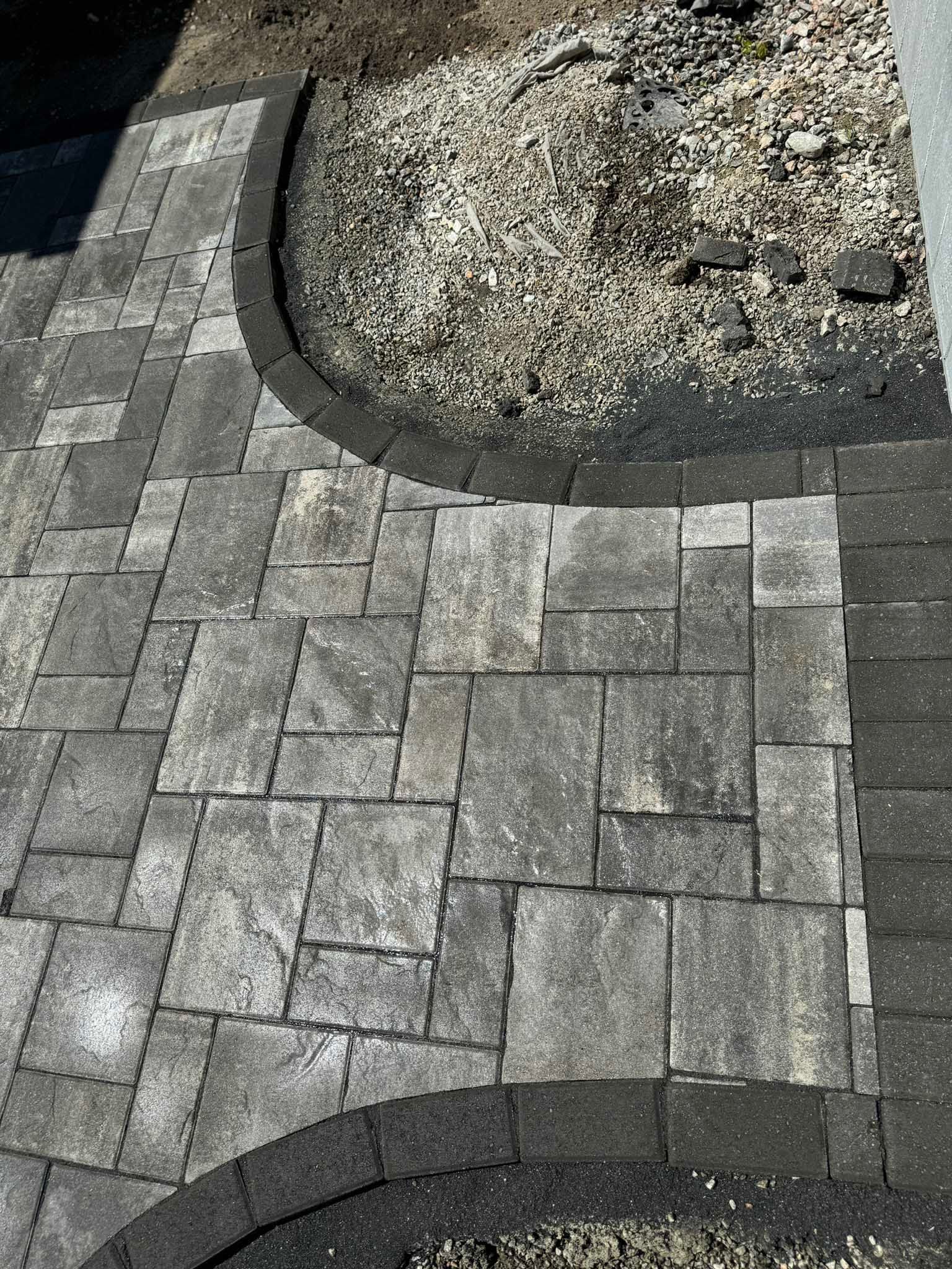 A textured gray paver patio with a dark border, curving around a patch of gravel and dirt.