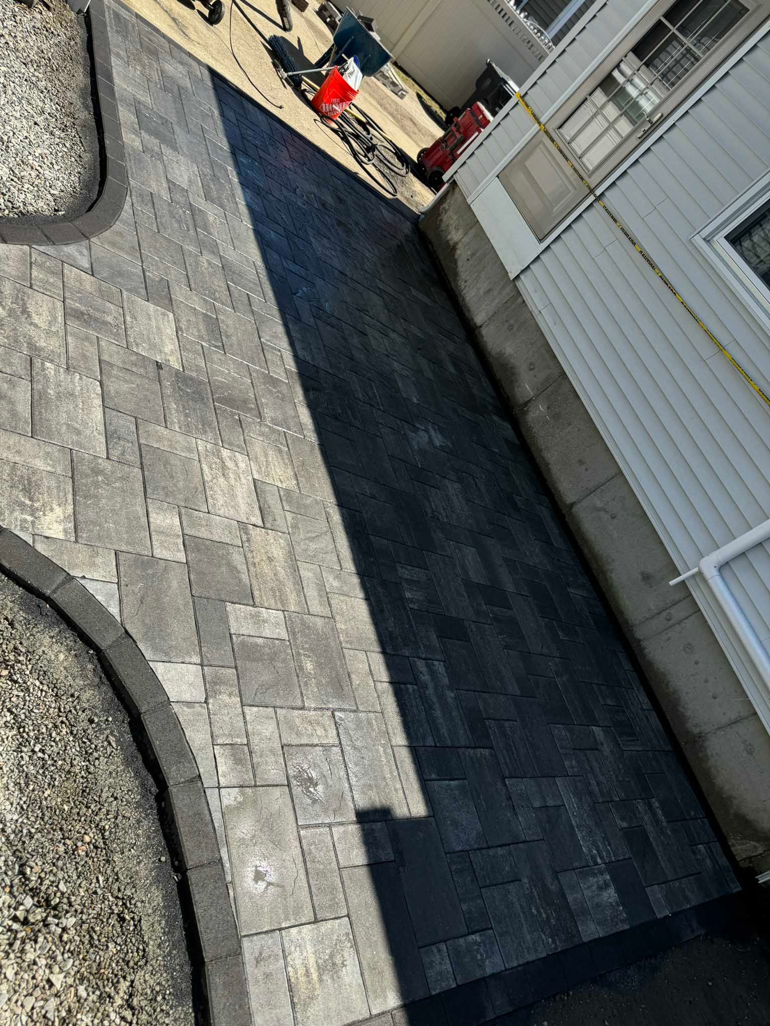 A paved stone walkway with a dark border runs alongside the light-colored exterior wall of a house.