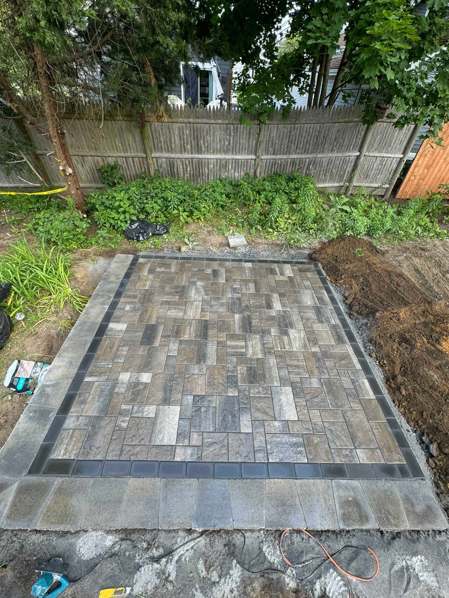 A rectangular stone patio featuring a mix of gray pavers surrounded by a darker border, set in a backyard near a fence.