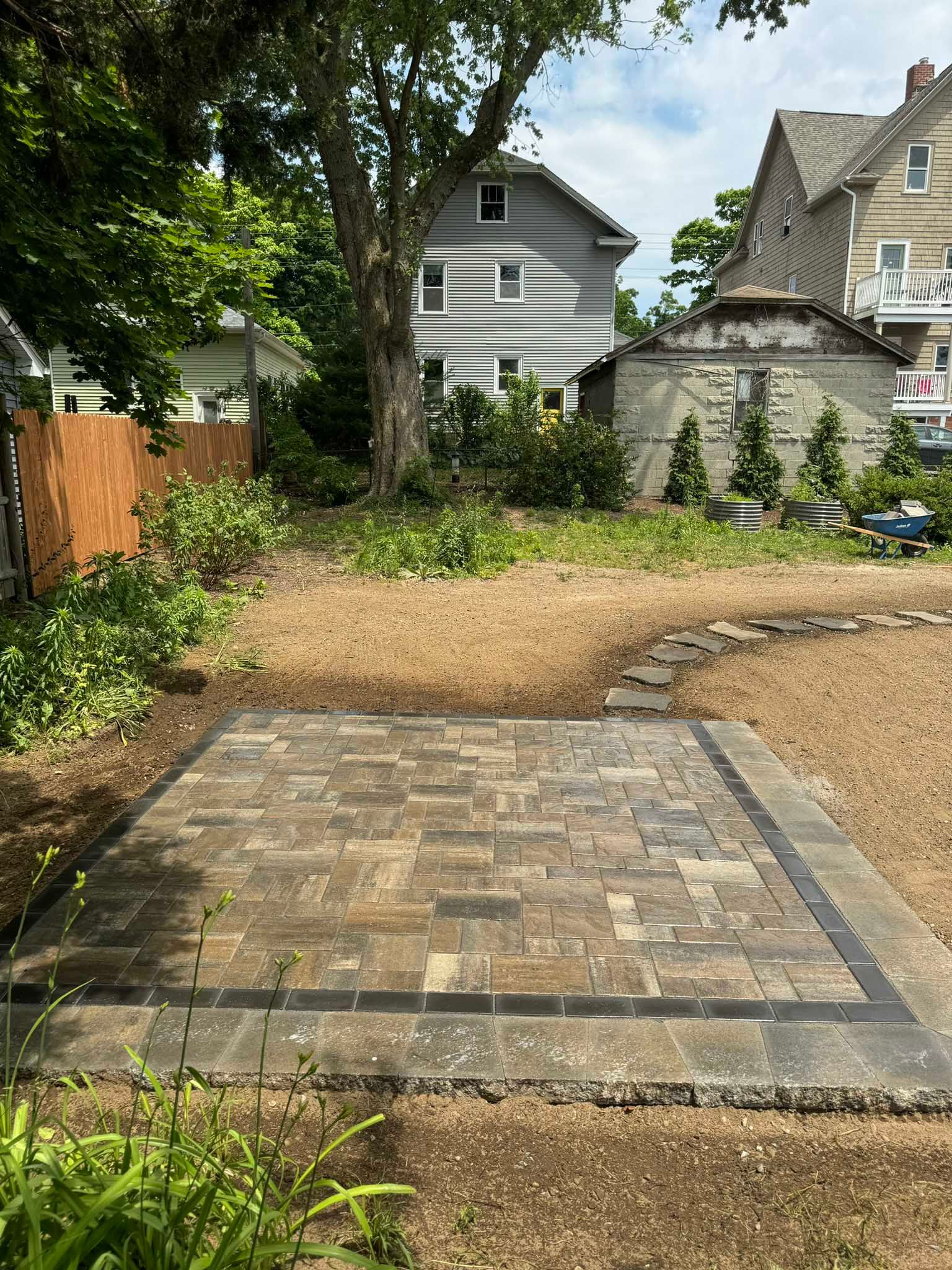 A rectangular stone paver patio sits in a backyard, bordered by a dark stone frame, with houses visible in the background.