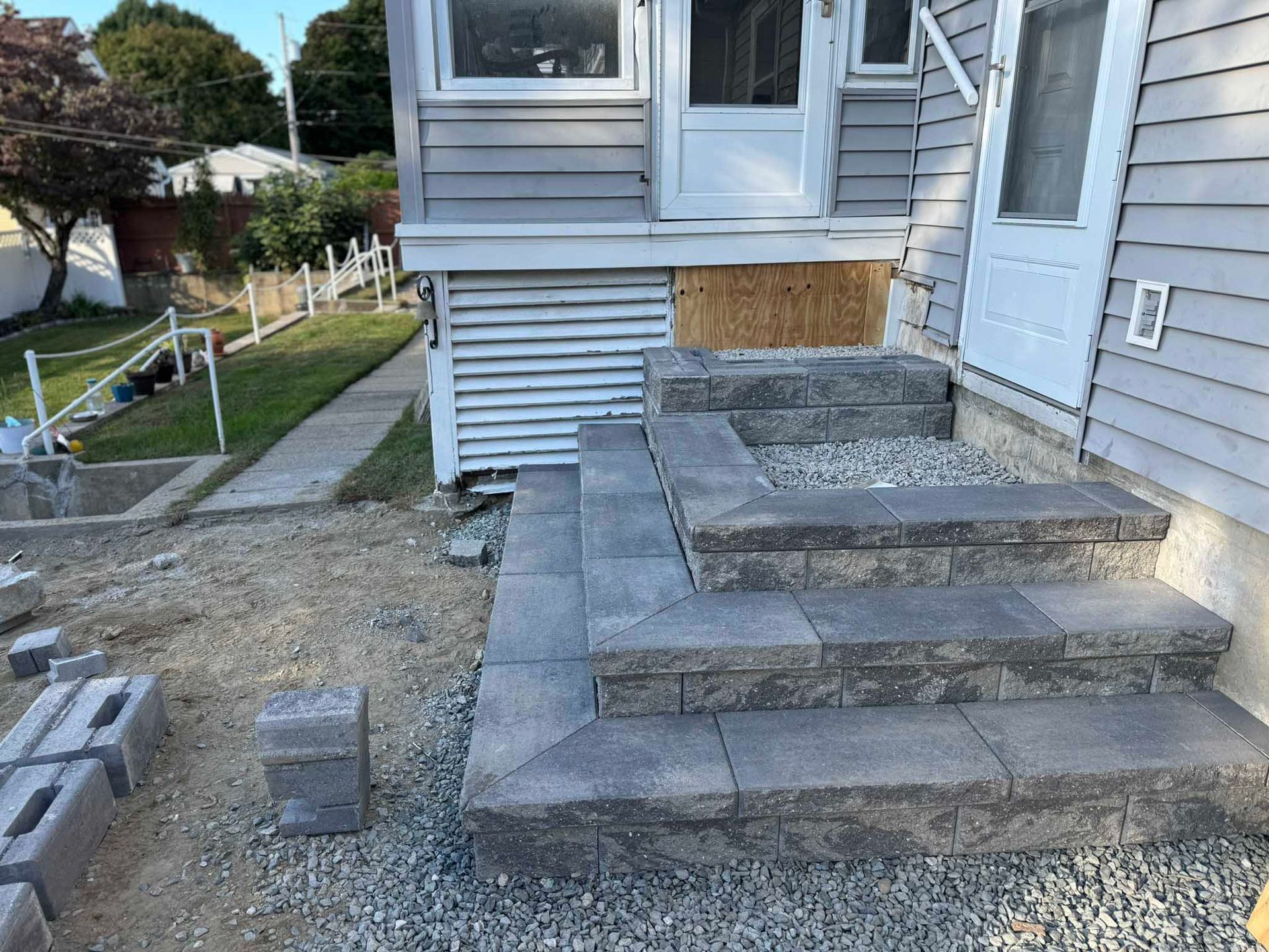 Newly constructed gray stone steps leading to a side door of a gray-sided house with a gravel ground surface.