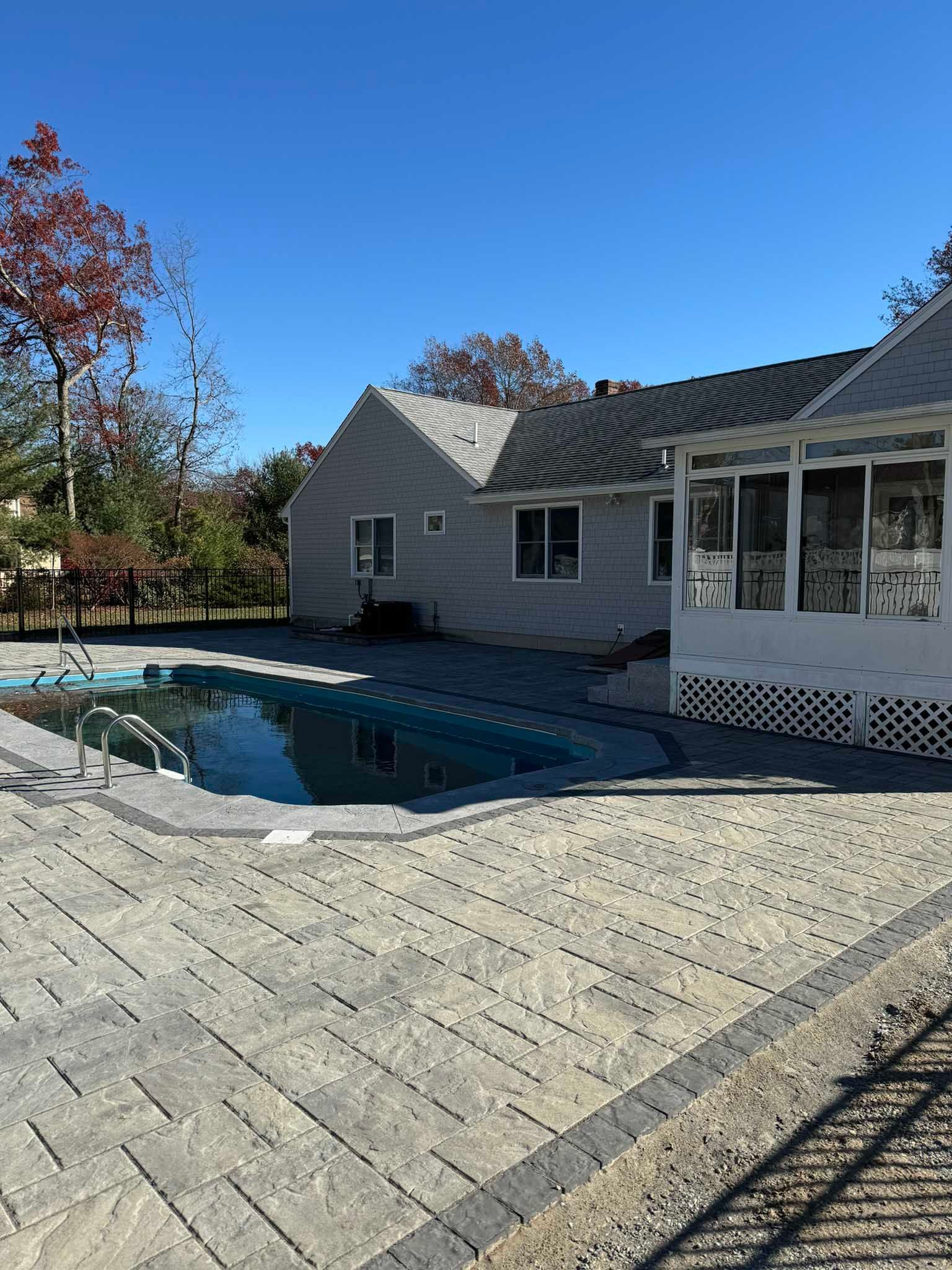 A backyard pool area with light stone pavers, a white sunroom, and a house under a clear blue sky.