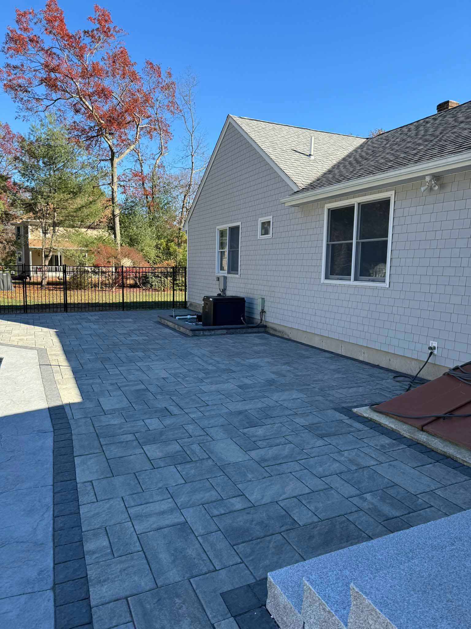 A backyard stone patio with a light gray border and inner dark gray paving stones extending from a light-sided house.