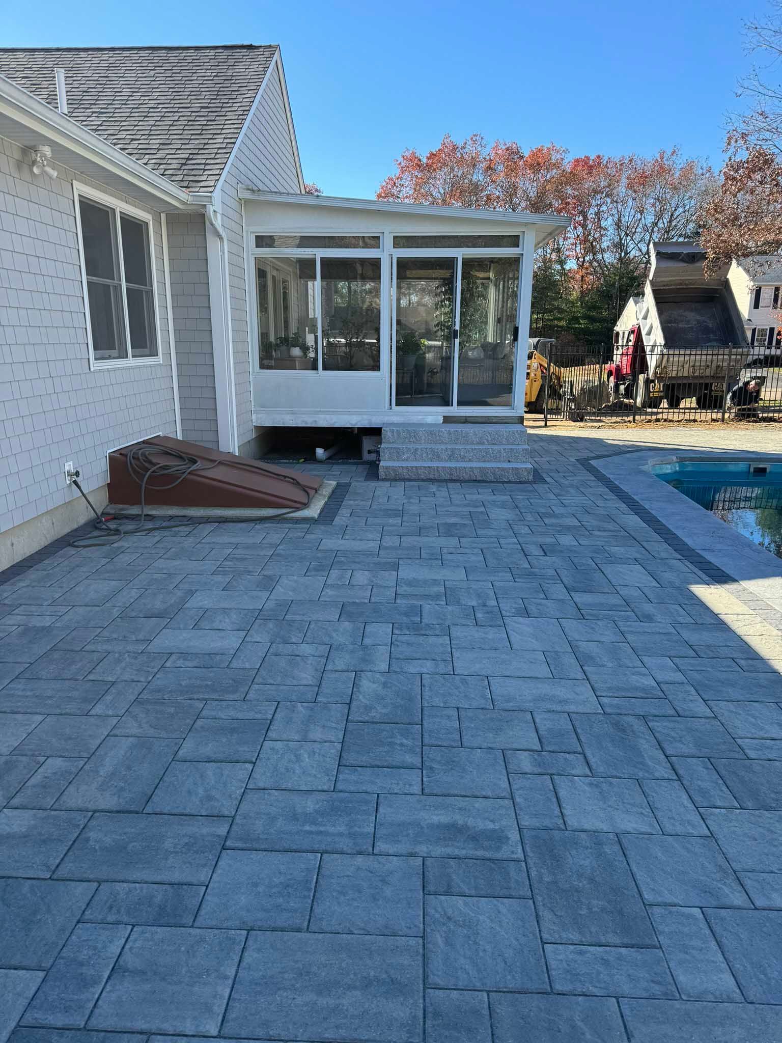 A modern backyard with a gray paver patio, a white sunroom, and a corner of a swimming pool under a bright blue sky.