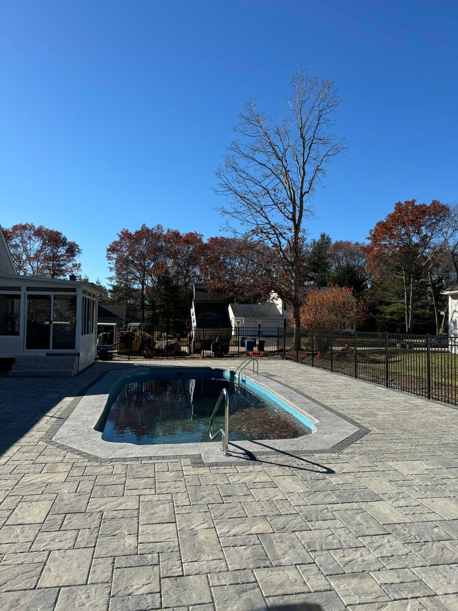 A rectangular swimming pool surrounded by gray stone pavers under a clear blue sky, with trees in the background.