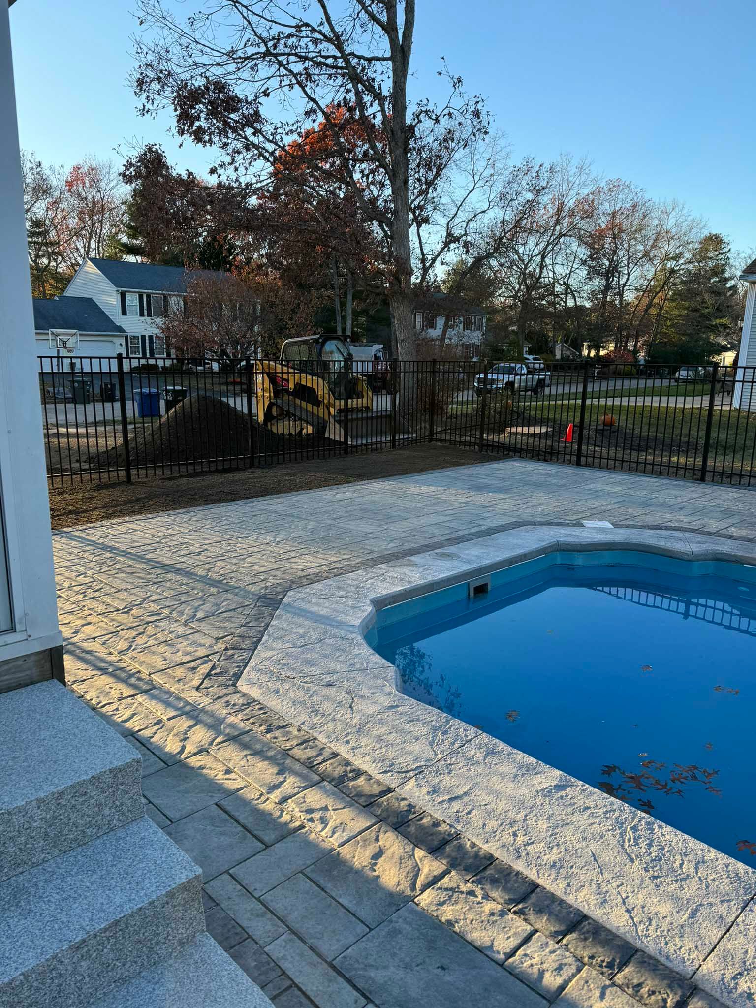 A square, blue swimming pool with textured stone coping, surrounded by a light-colored paver patio under a clear blue sky.