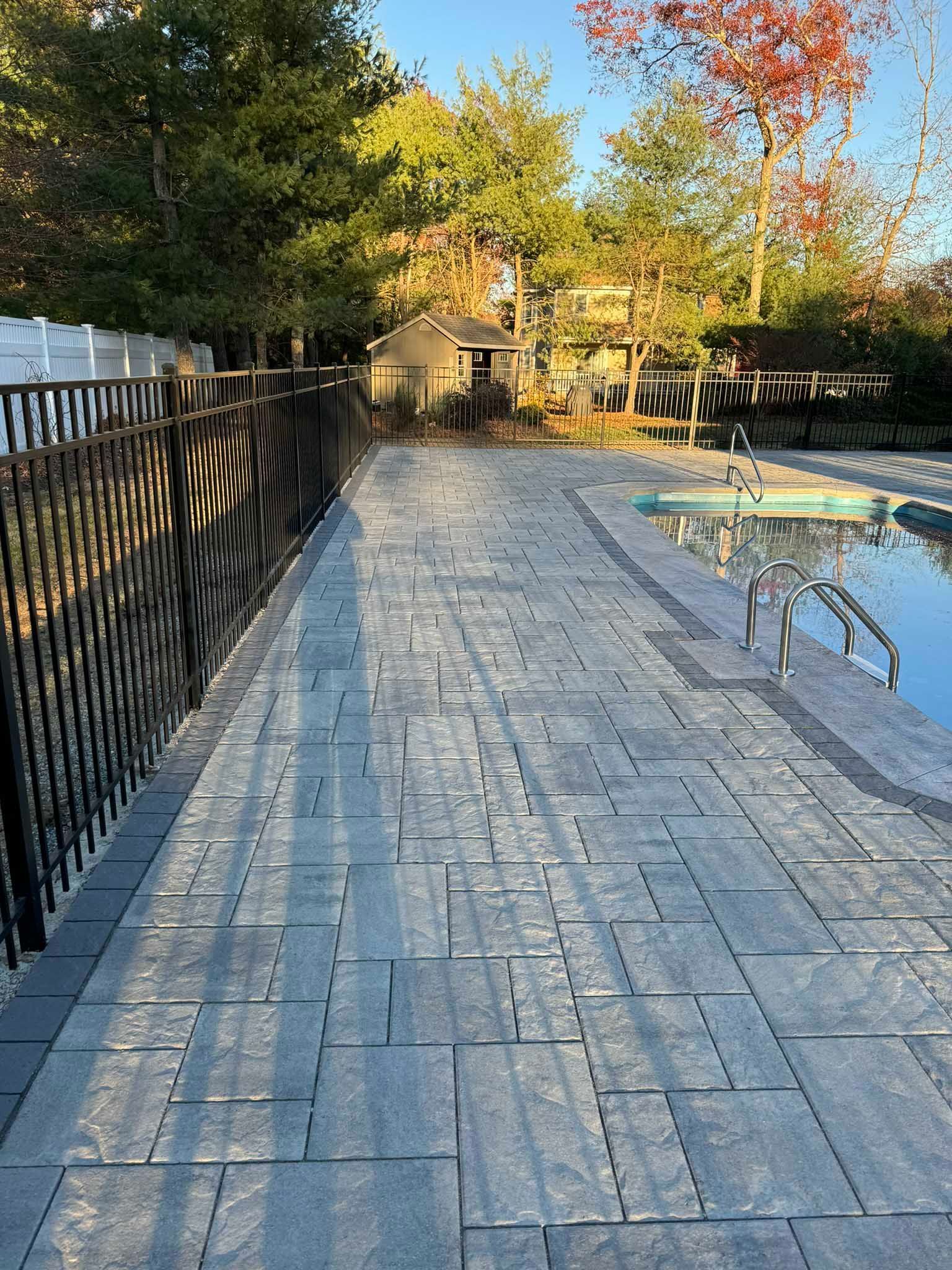 A patio made of gray rectangular pavers runs alongside a swimming pool, bordered by a dark metal fence and trees.