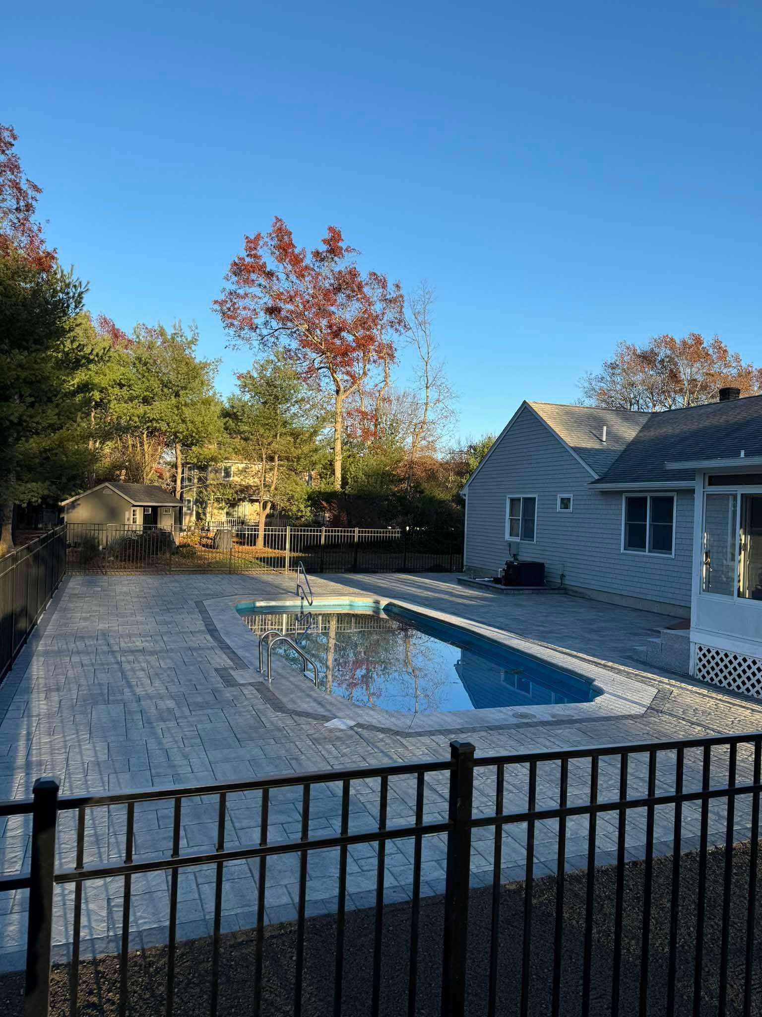A backyard featuring a rectangular swimming pool surrounded by a large gray paver patio and a black metal fence.