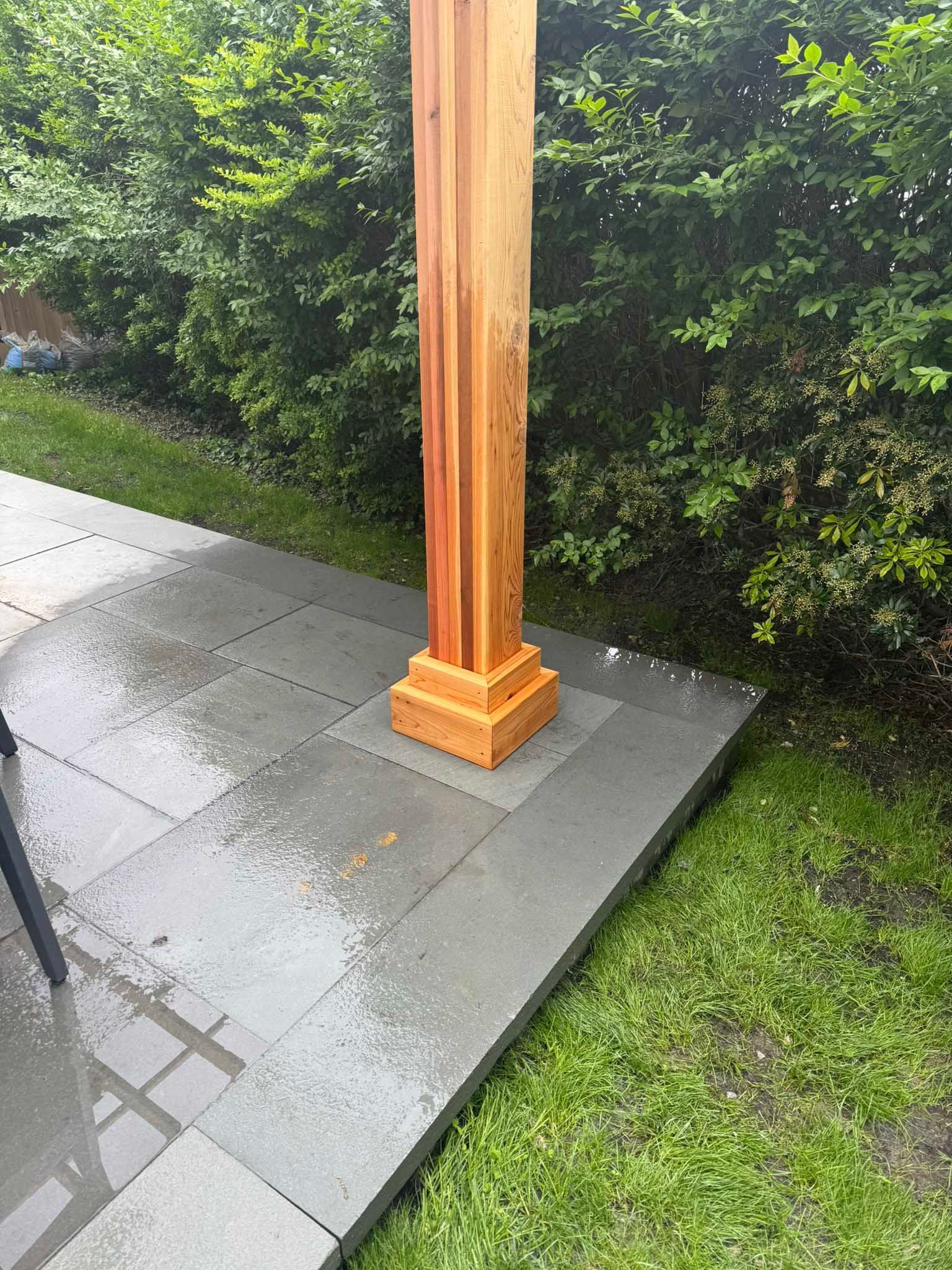 A wooden pergola post with a decorative base, installed on a gray stone patio beside a green hedge.
