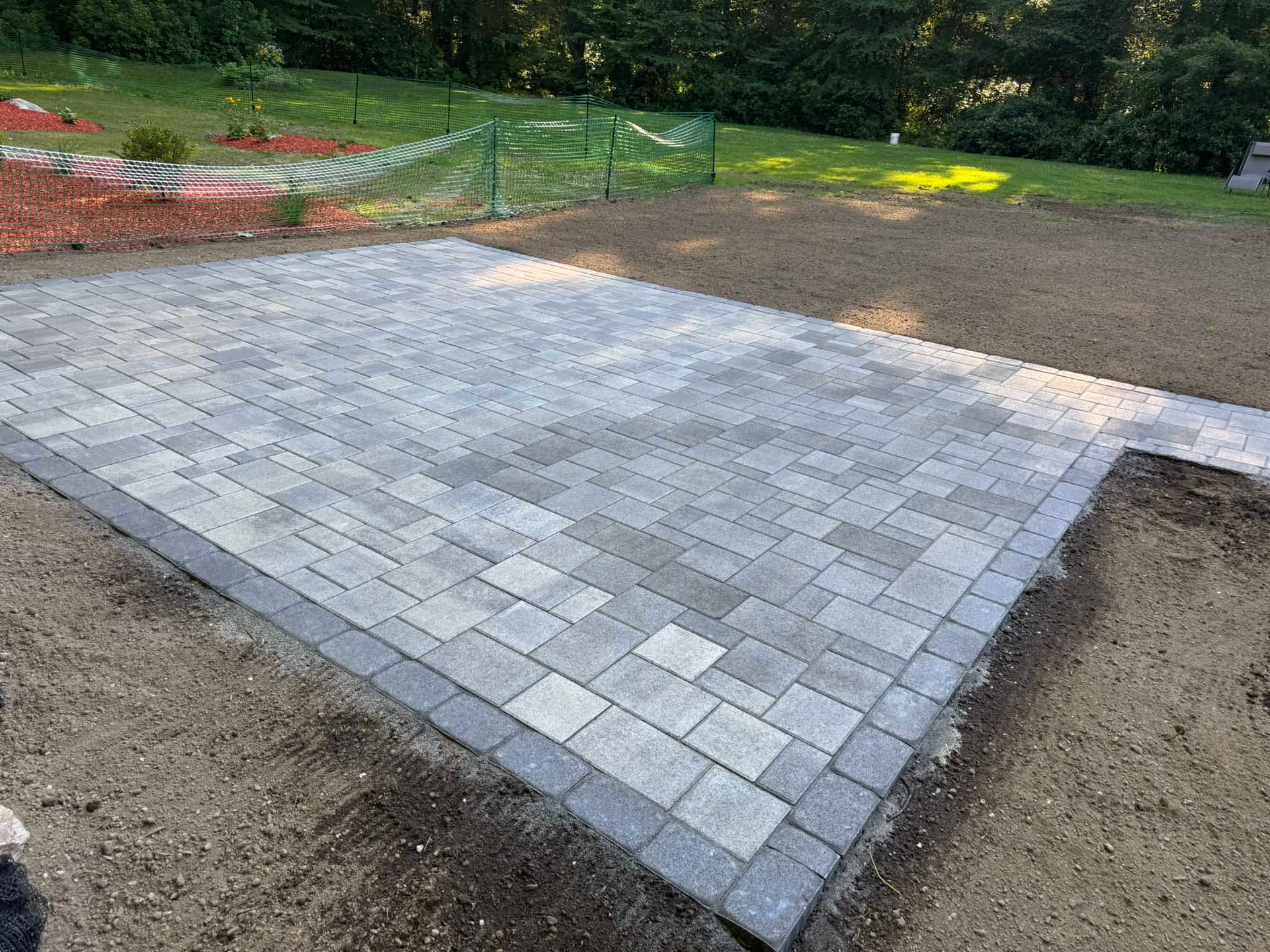 A newly installed rectangular paver patio with a dark border, surrounded by unfinished dirt ground and grass.