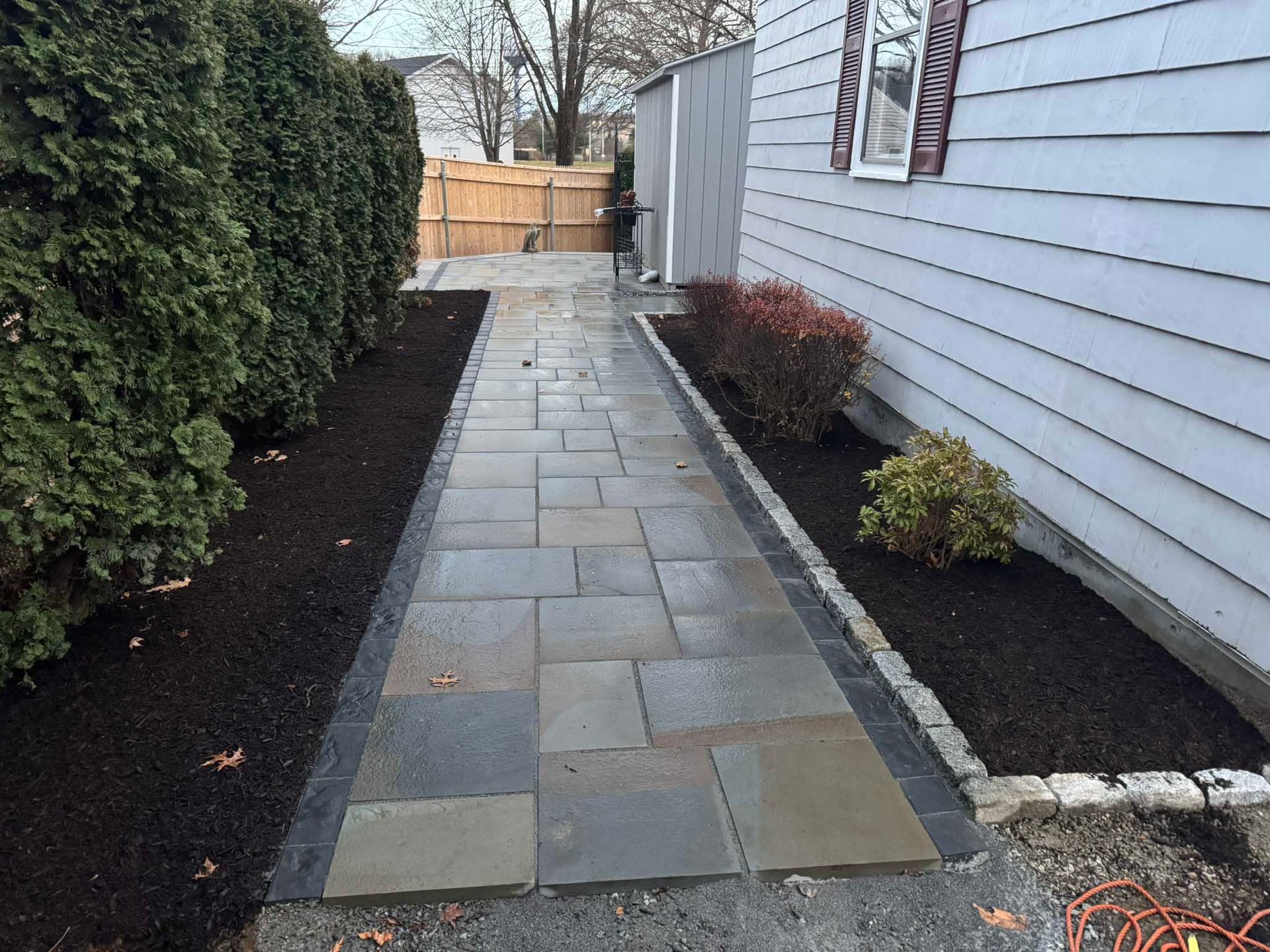 A stone walkway with a dark border leads toward a house with white siding, bordered by mulch, bushes, and a hedge.