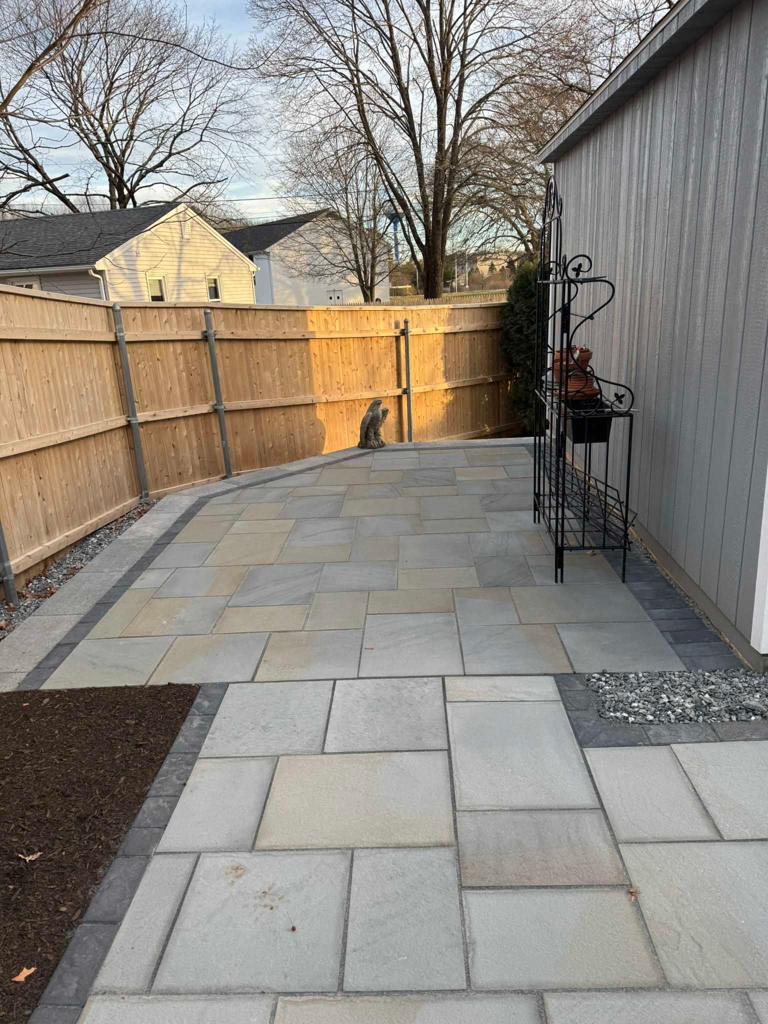 A stone patio with a dark border, adjacent to a house wall and a wooden fence in a backyard setting.