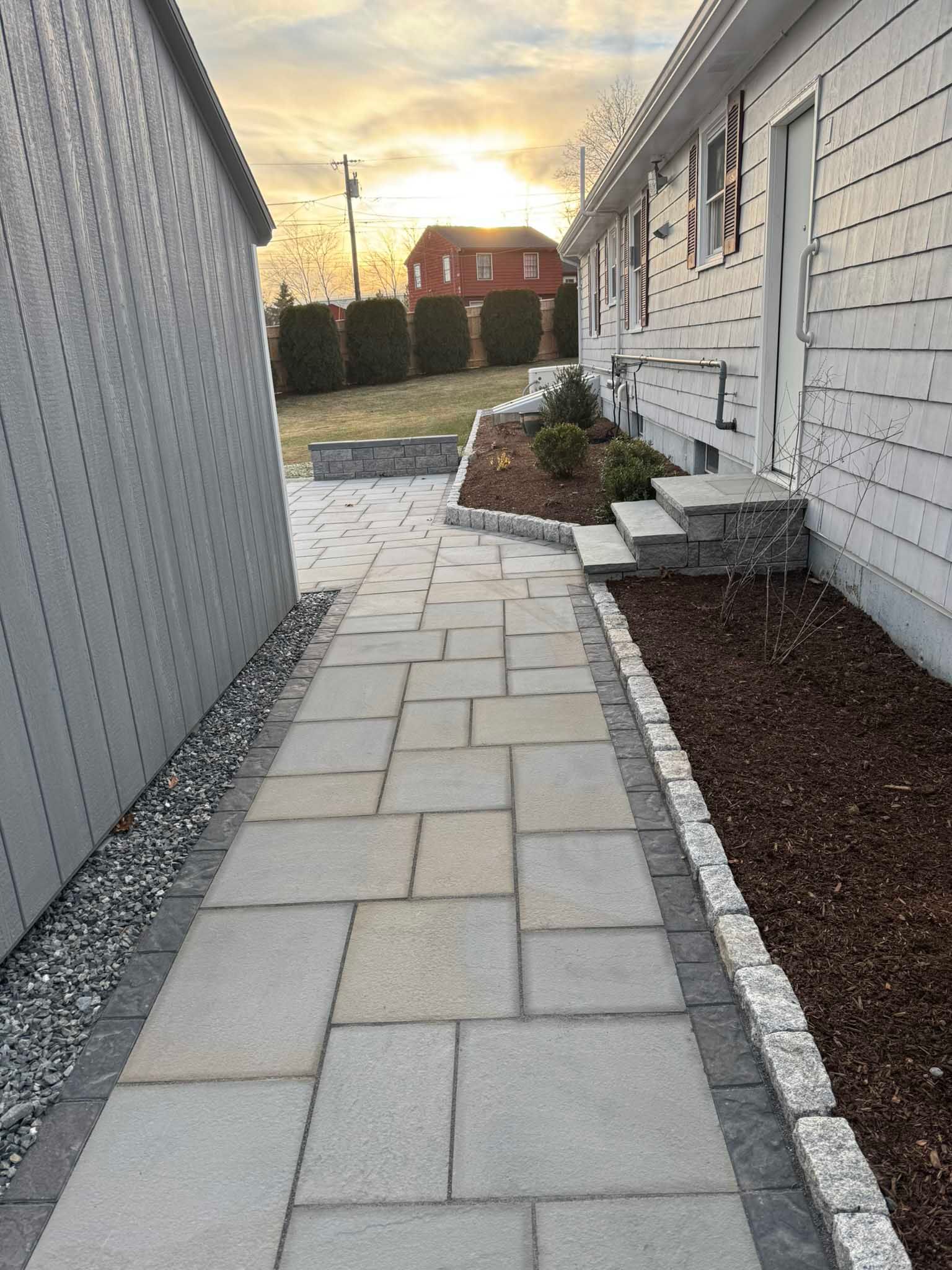 A paved stone walkway leads past a white siding house toward a backyard at sunset, with a mulched garden bed to the right.
