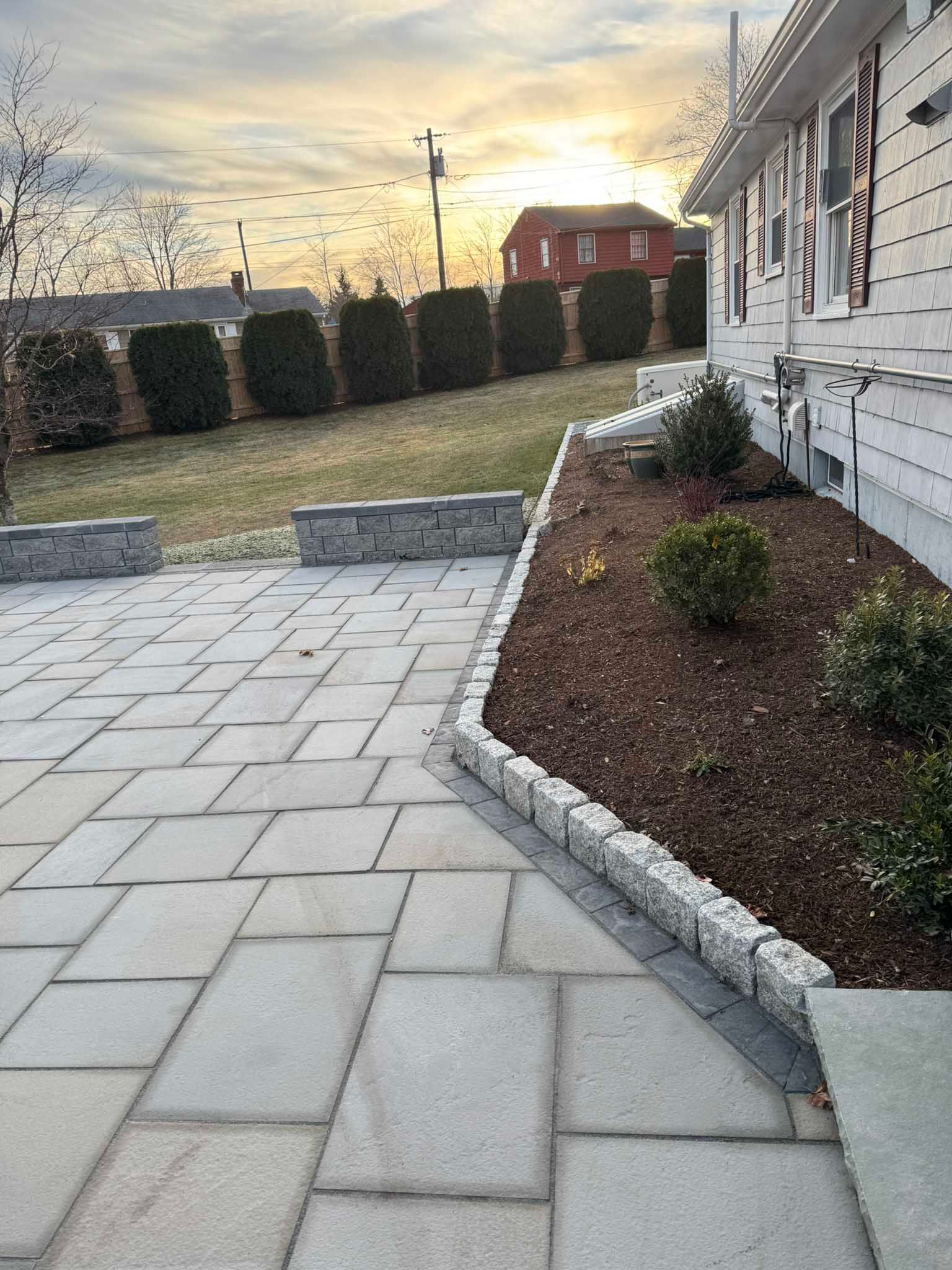 A stone patio with a garden bed featuring a manicured hedge and a white house exterior at sunset.