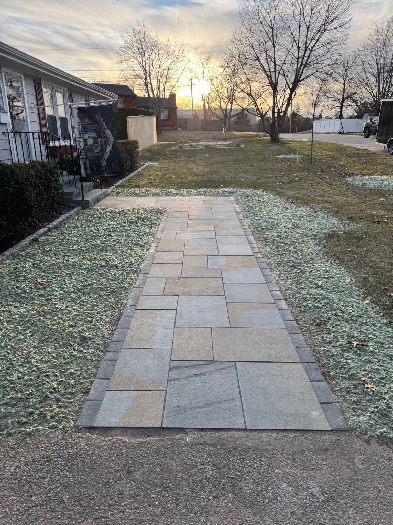 A flagstone walkway with a dark brick border runs through a front yard toward a house at sunset.
