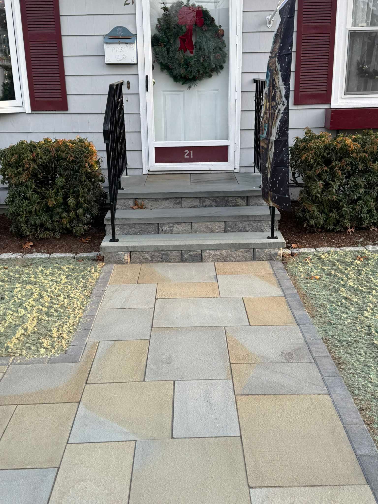 A front entryway with stone steps, a paved walkway, maroon shutters, a green wreath on the door, and two small bushes.
