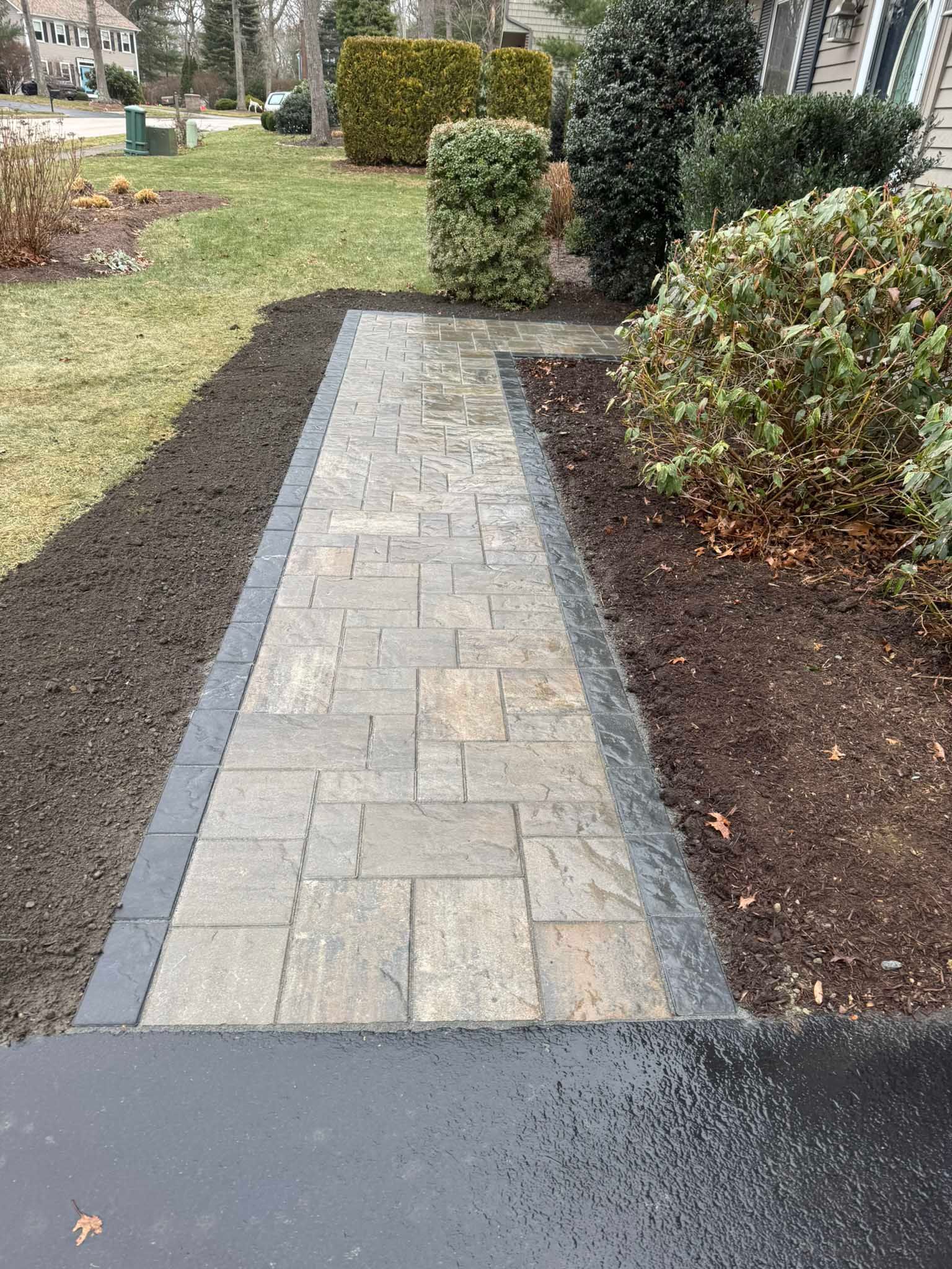 A paved walkway with a dark border leads from a driveway toward a house, flanked by dark mulch and evergreen shrubs.