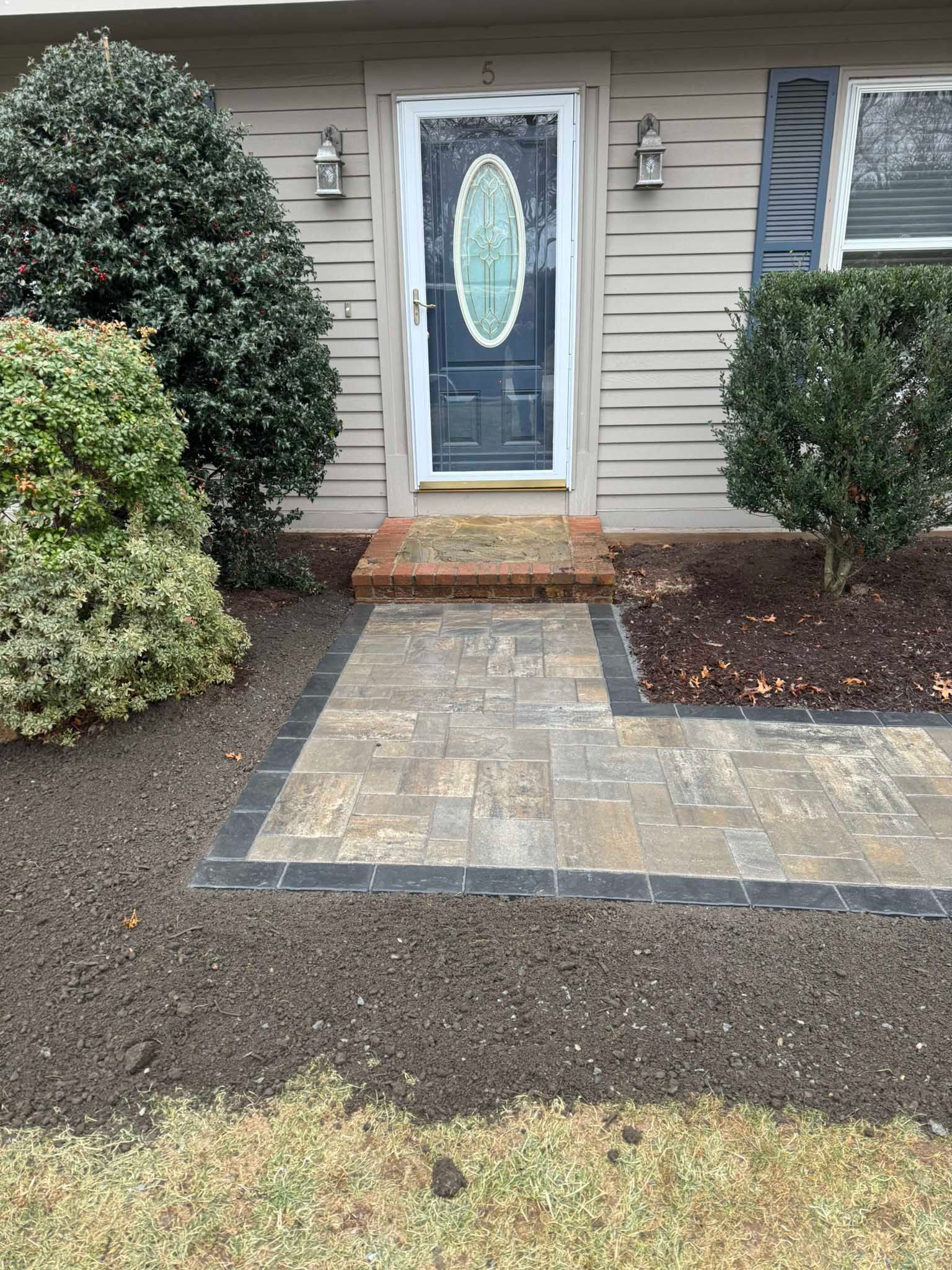 A front entryway with a dark blue door, two large shrubs, and a paved walkway leading to brick steps.