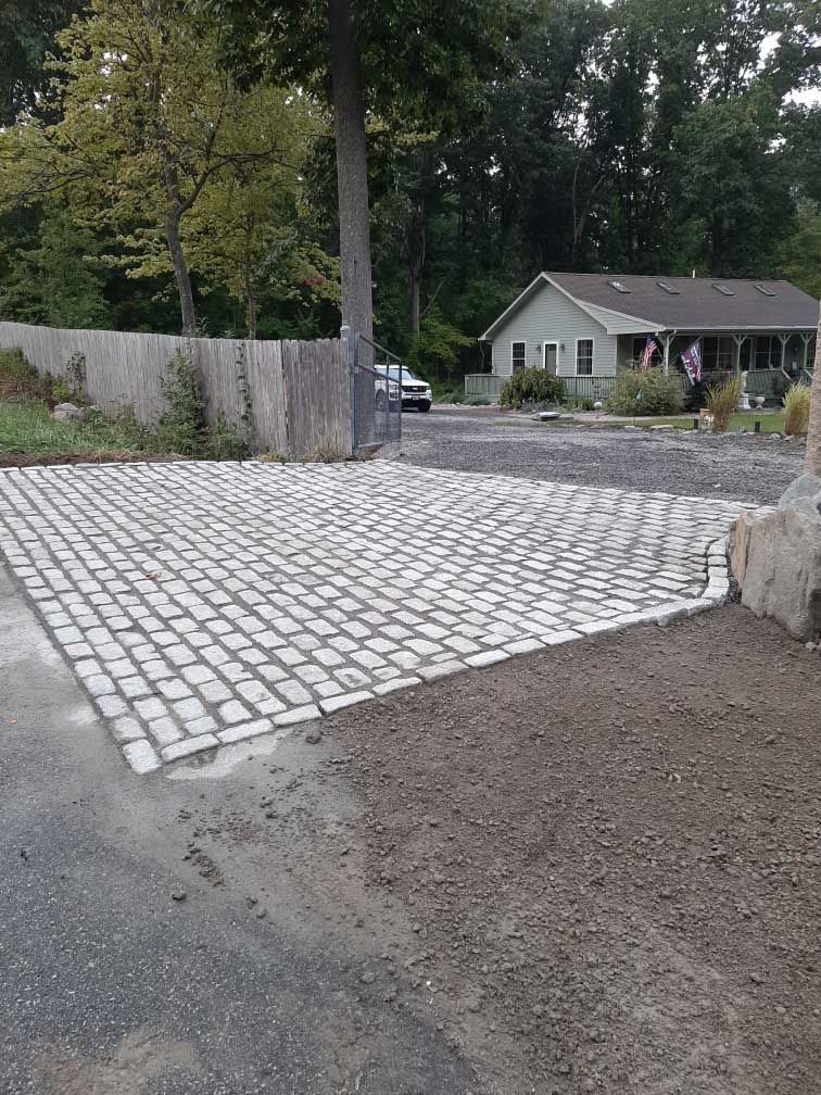 A rectangular patch of light gray cobblestones sits at the entrance of a gravel driveway next to a grassy lawn and fence.