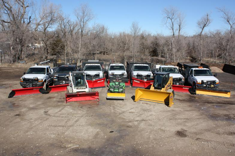 Snow removal vehicles lined up outside. Several trucks with plows and two pieces of heavy machinery. Blue sky and trees in the background.