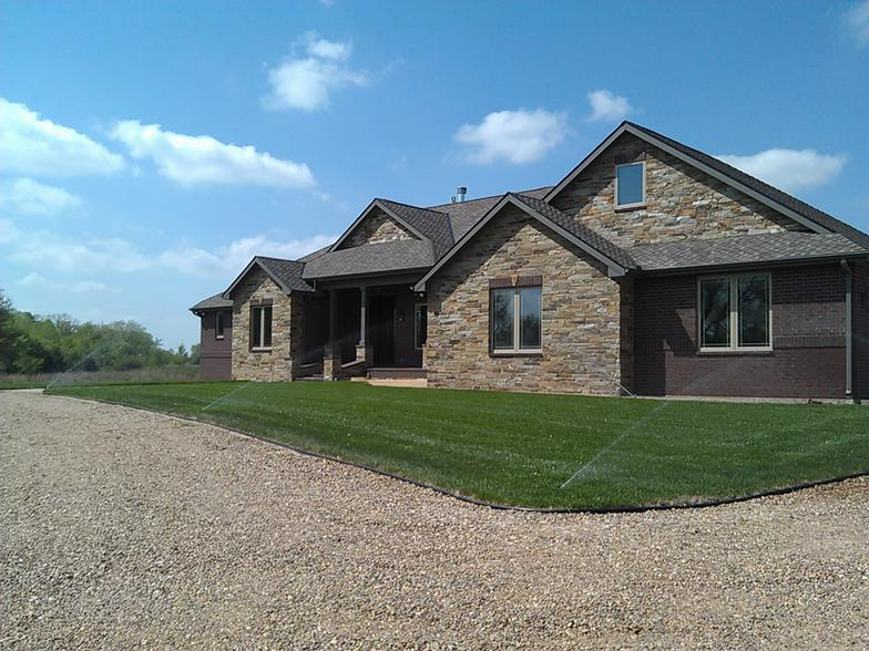 Stone and wood home with green lawn and gravel driveway under a blue sky.