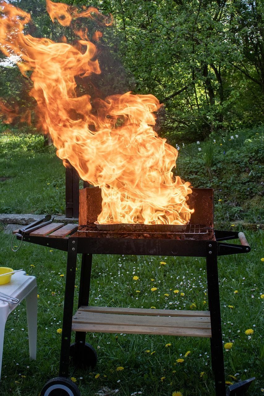 Large grill with a tall flame burning outdoors in a green yard, with a table nearby