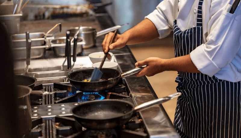 Chef stirring food in pans on a busy commercial stovetop with blue gas flames