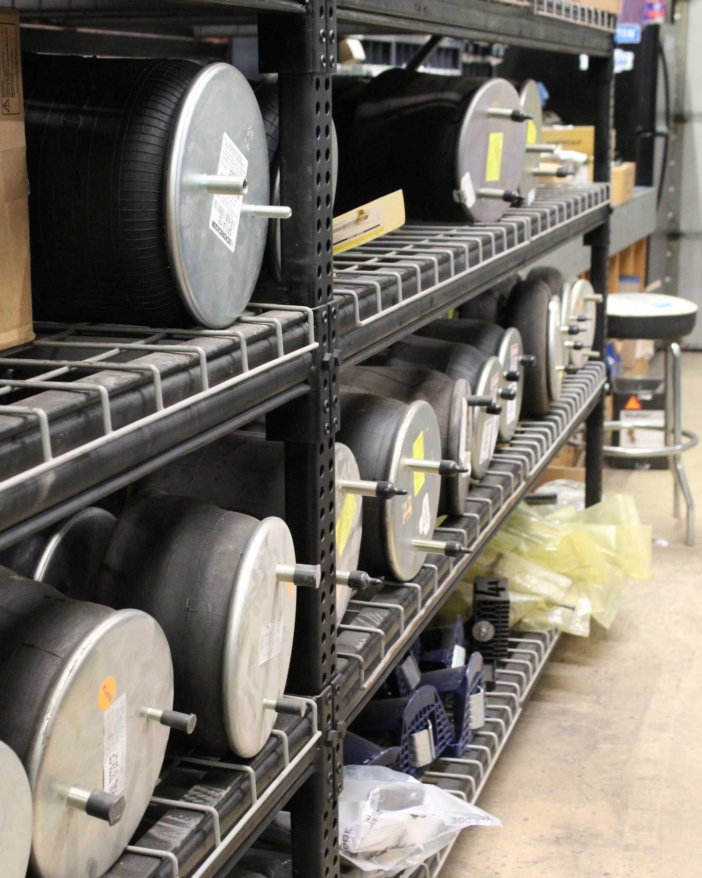 Black industrial shelves stacked with rows of cylindrical, black air springs with metal end plates in a warehouse.