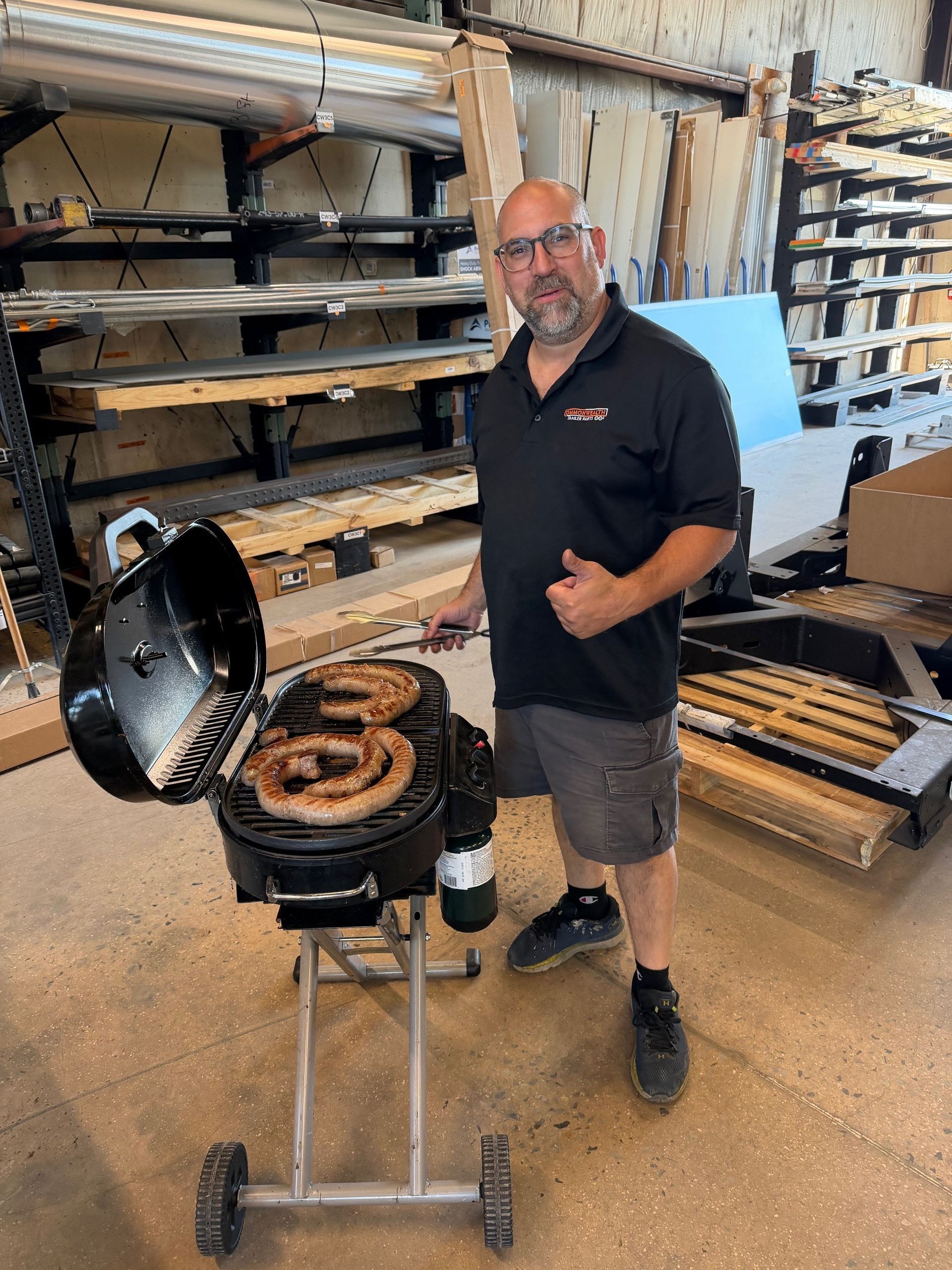Man grilling at a shop display with a black kettle grill, stacked coiled meat, and trays of food behind him