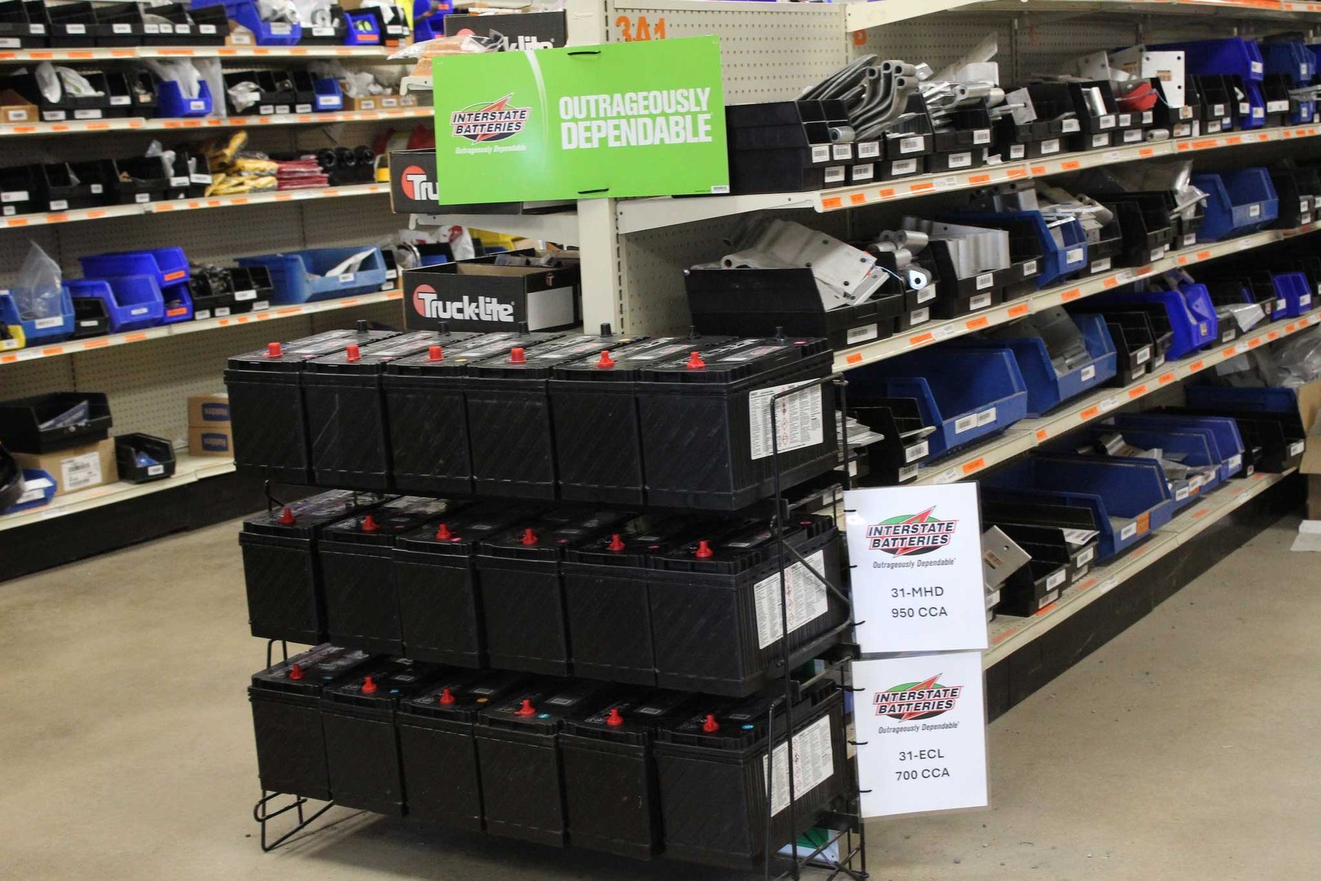 A three-tier shelf filled with black lead-acid batteries in an automotive parts store, near aisle shelves with blue bins.