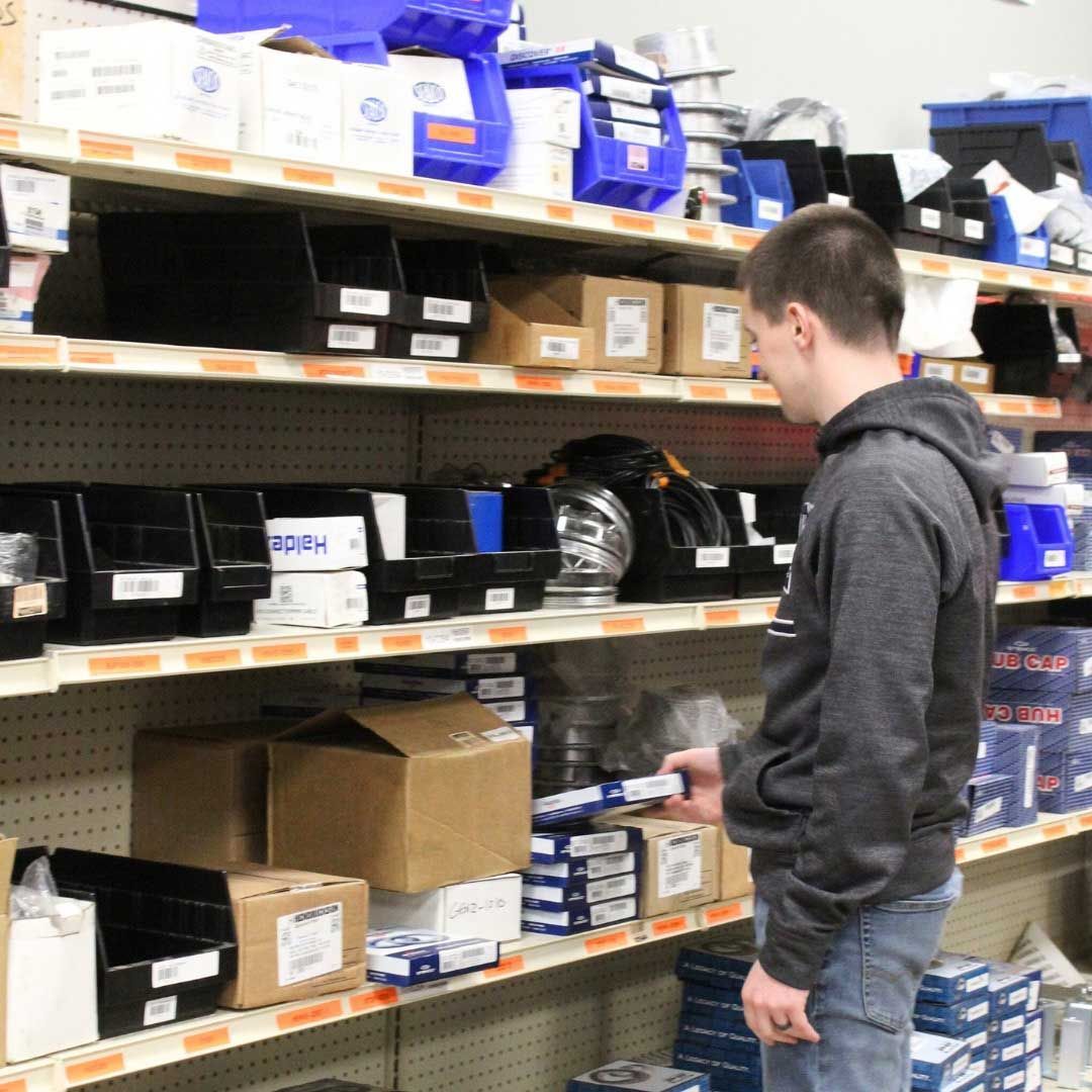 A person in a gray hoodie browses shelves of organized hardware and boxed inventory in a supply store.