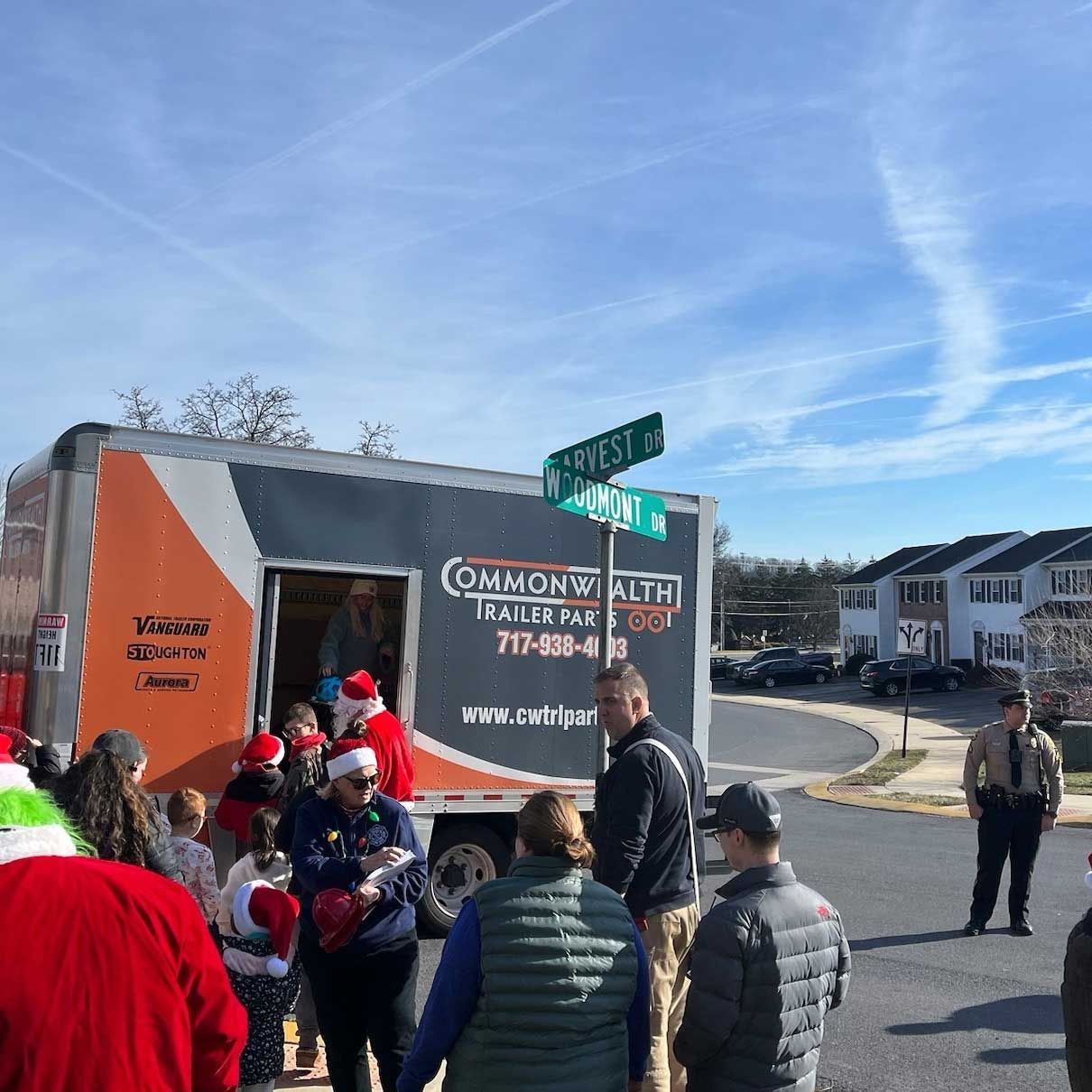 People gather near a Commonwealth Trailer rental truck decorated for Christmas at a street intersection on a sunny day.