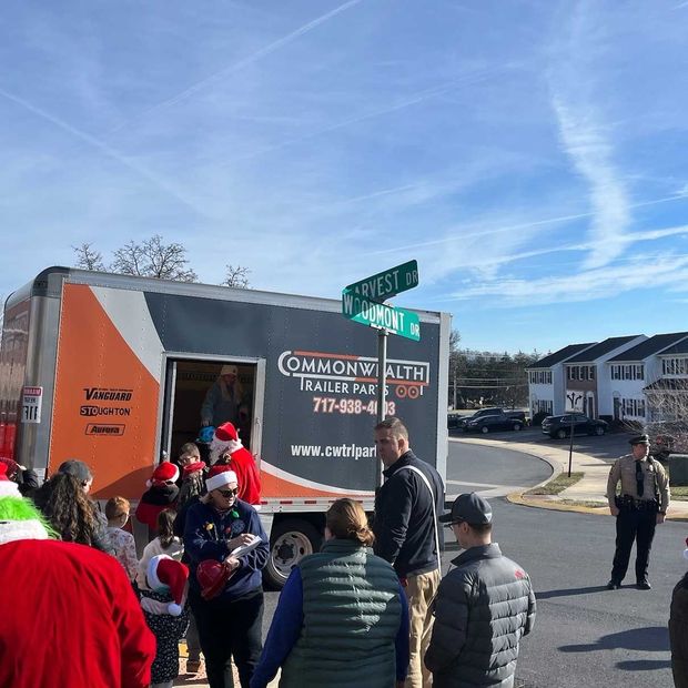 People gather near a Commonwealth Trailer rental truck decorated for Christmas at a street intersection on a sunny day.