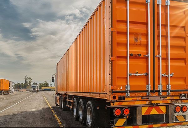 A bright orange shipping container on a semi-truck trailer parked on an asphalt lot under a cloudy sky.
