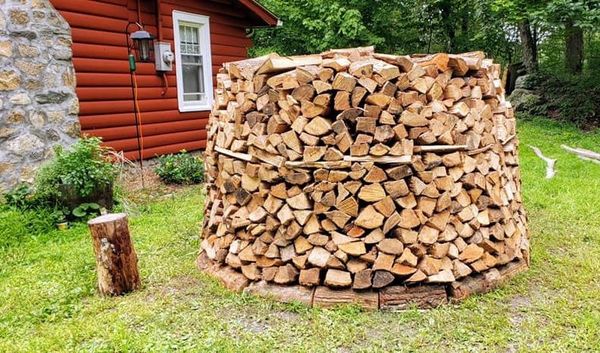 Stacked firewood beside a red log cabin and stone wall in a grassy yard