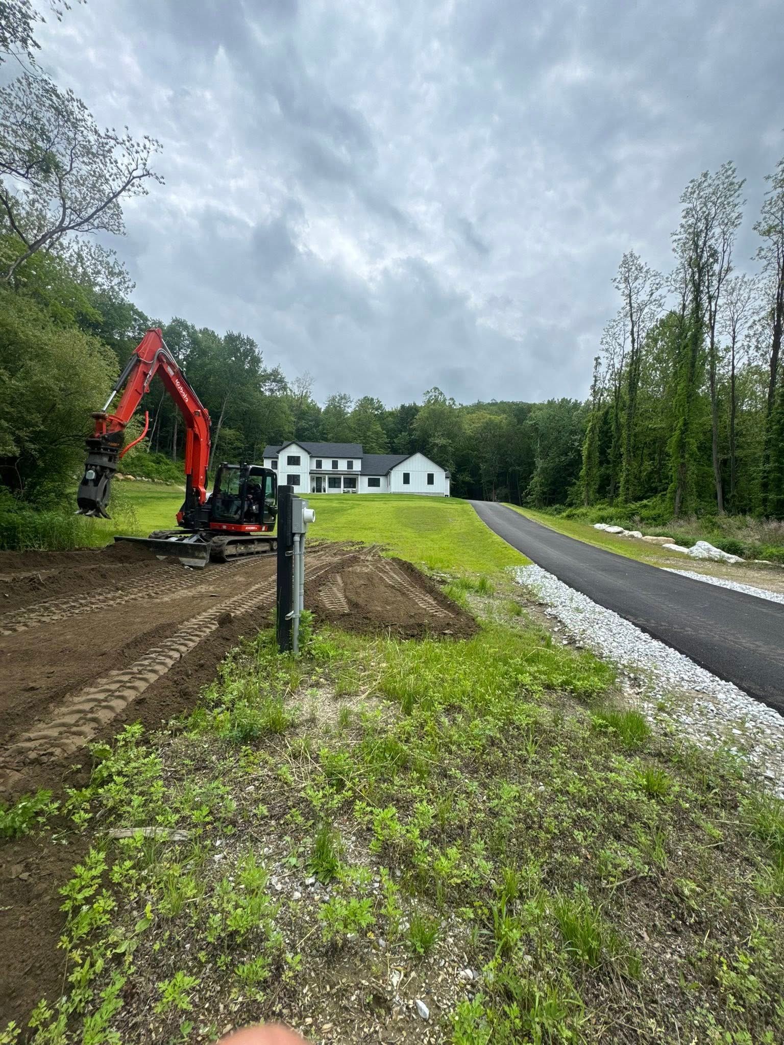 Excavator beside a gravel road under construction, with a white house and cloudy sky in the background