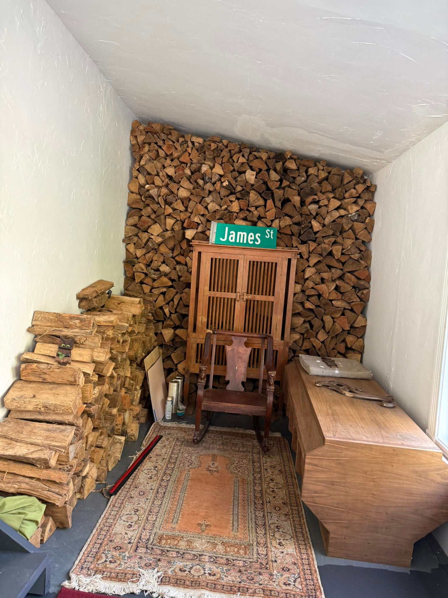 Wood-paneled room with stacked firewood, patterned rug, and a wooden rocking chair under a small green sign.